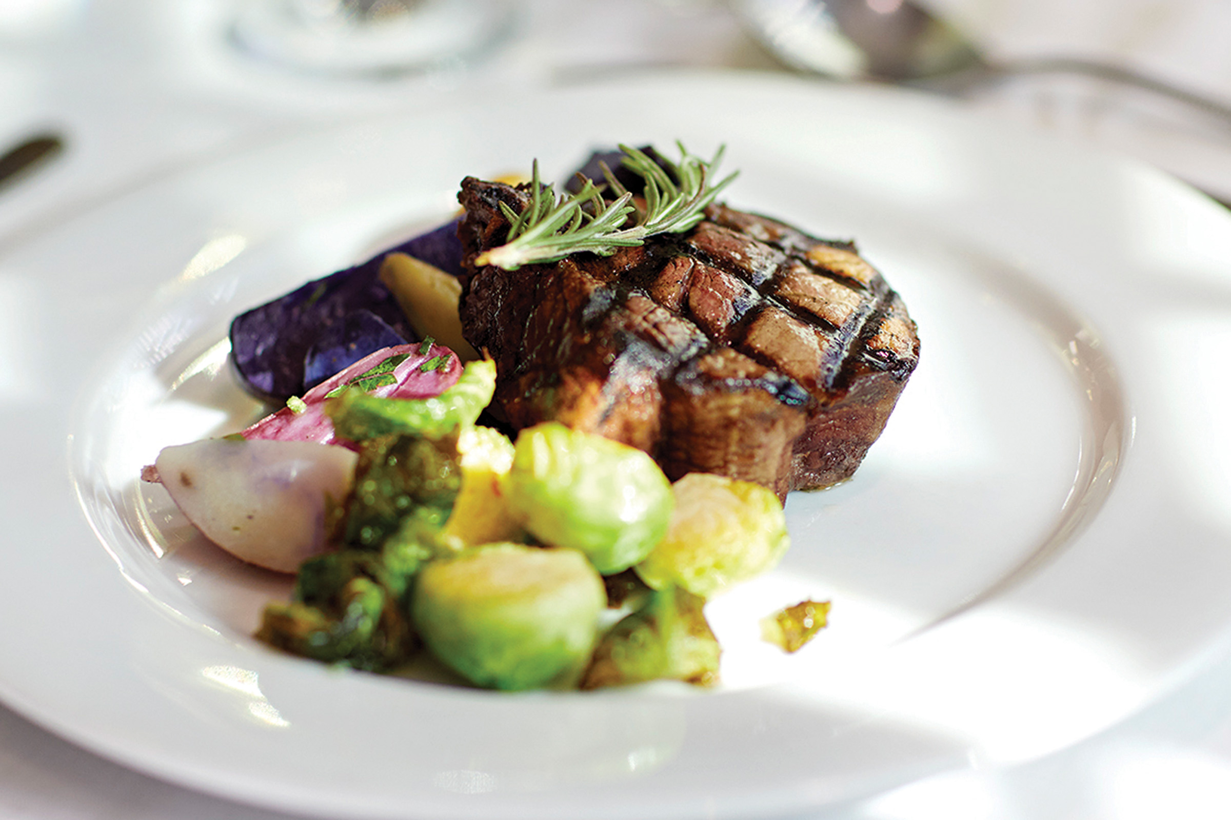 A grilled steak garnished with a sprig of rosemary, served with roasted Brussels sprouts, purple potatoes, and other vegetables on a white plate.