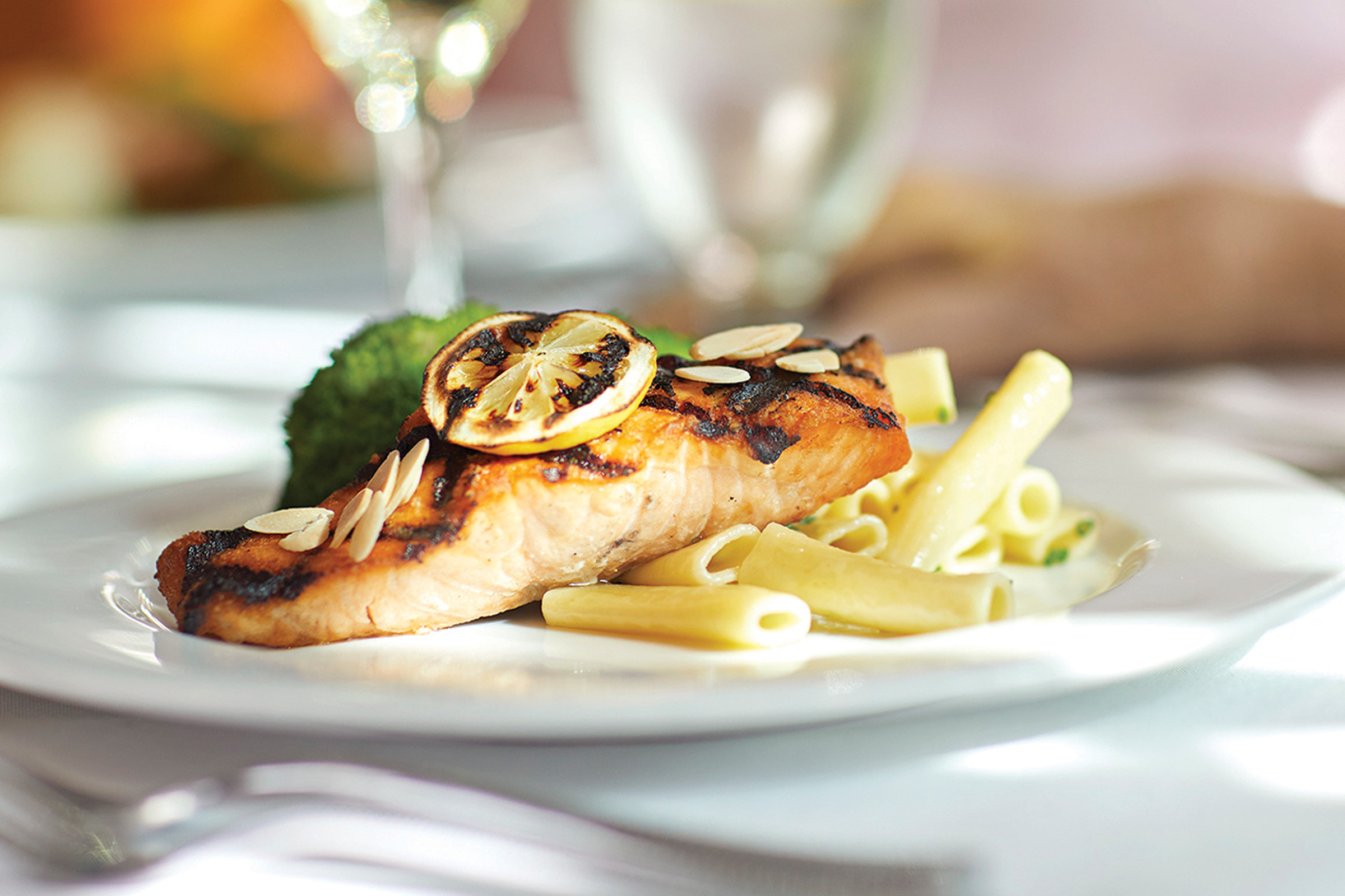 A plated meal featuring a grilled salmon fillet topped with grilled lemon slices and almond slivers, served with penne pasta in a creamy sauce, on a white plate with a blurred background.