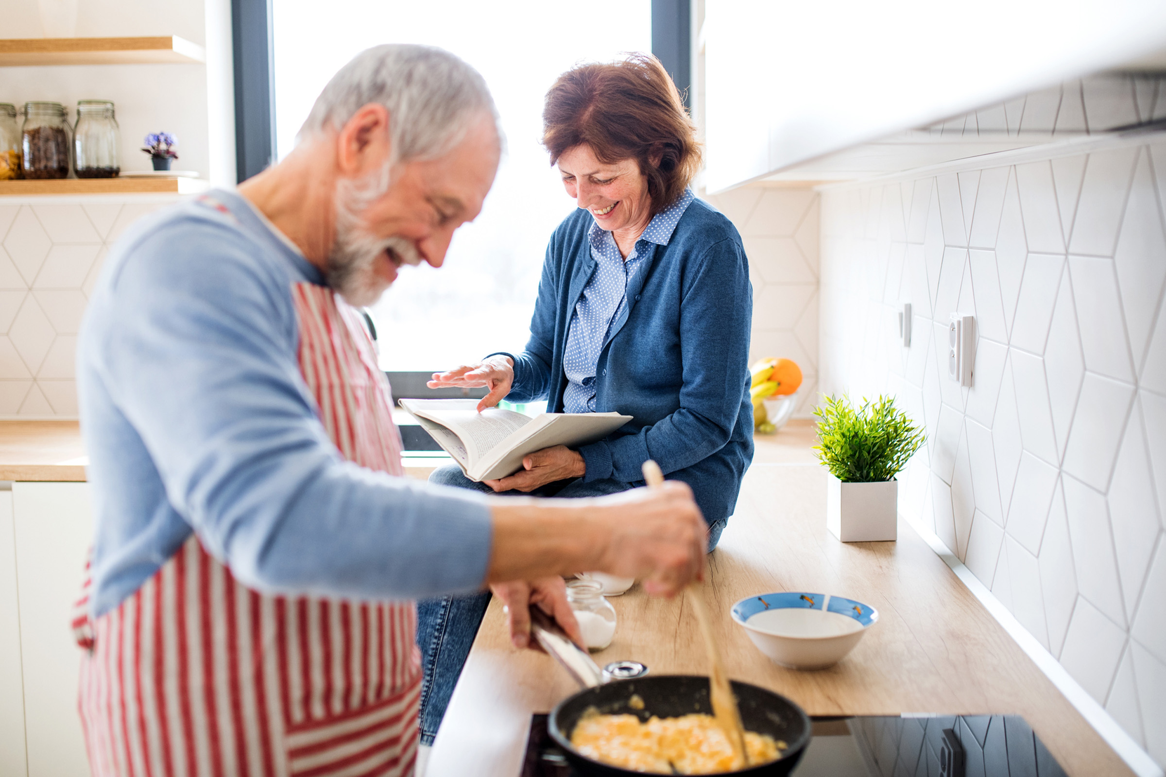 An older man in an apron cooks eggs in a pan while smiling. A woman, sitting on the kitchen counter, reads a book and looks at him, both appearing happy and relaxed in a modern, bright kitchen.