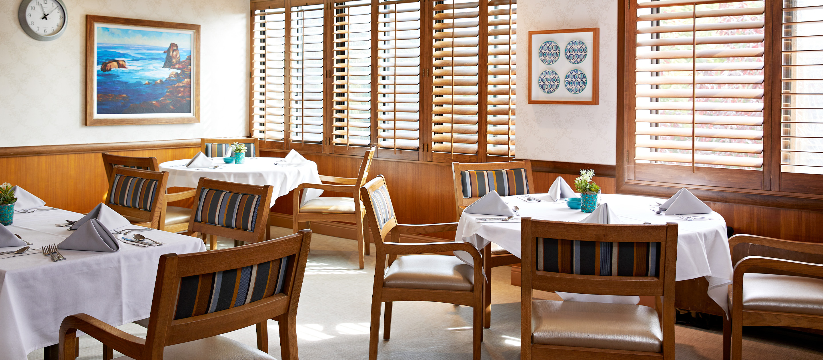 A bright restaurant dining area with wooden chairs, tables covered in white cloths, folded napkins, wall art, a clock, and sunlight streaming through wooden blinds on the windows.
