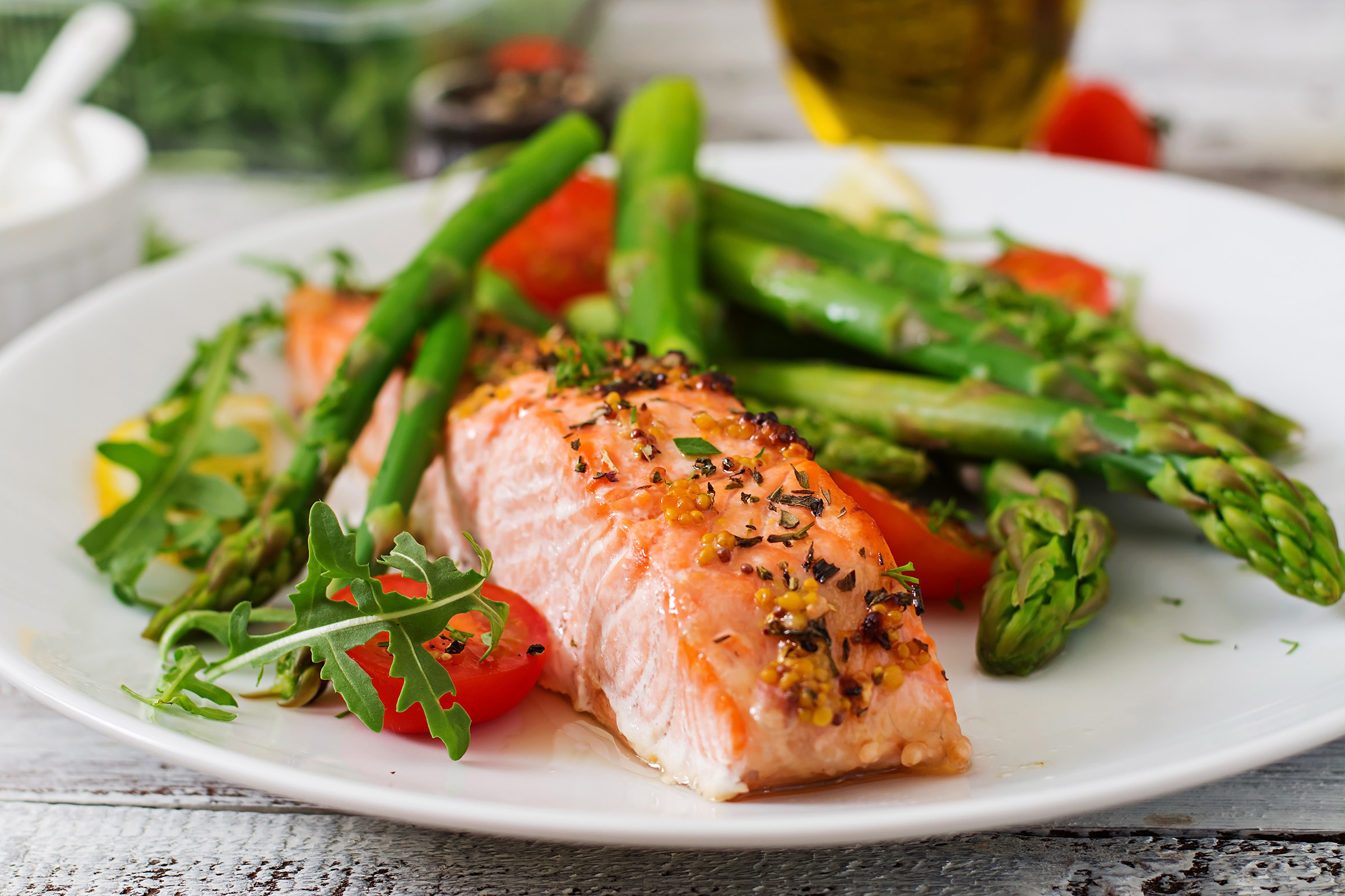 A plate with a cooked salmon fillet topped with herbs, served with asparagus, cherry tomatoes, arugula, and lemon slices on a white dish.