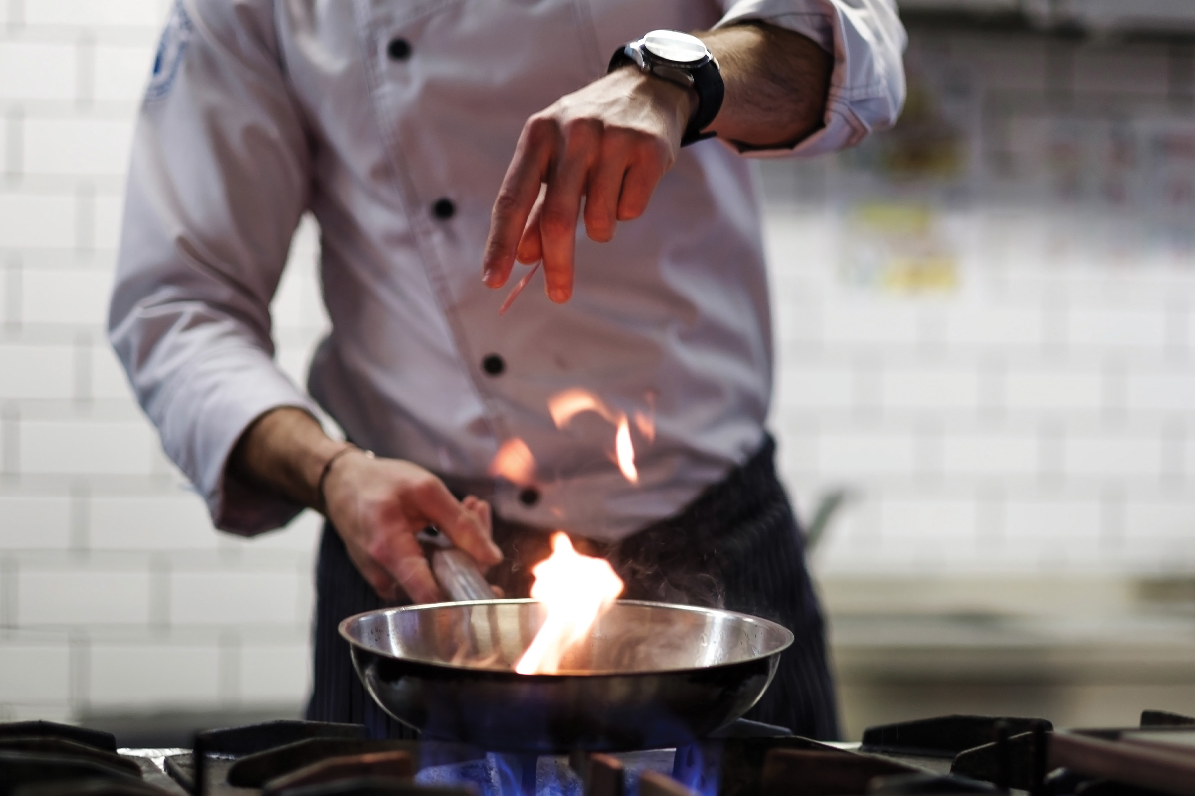 A chef in a white uniform sprinkles seasoning into a flaming pan on a stove, with blue flames visible underneath, in a professional kitchen with white tiled walls.