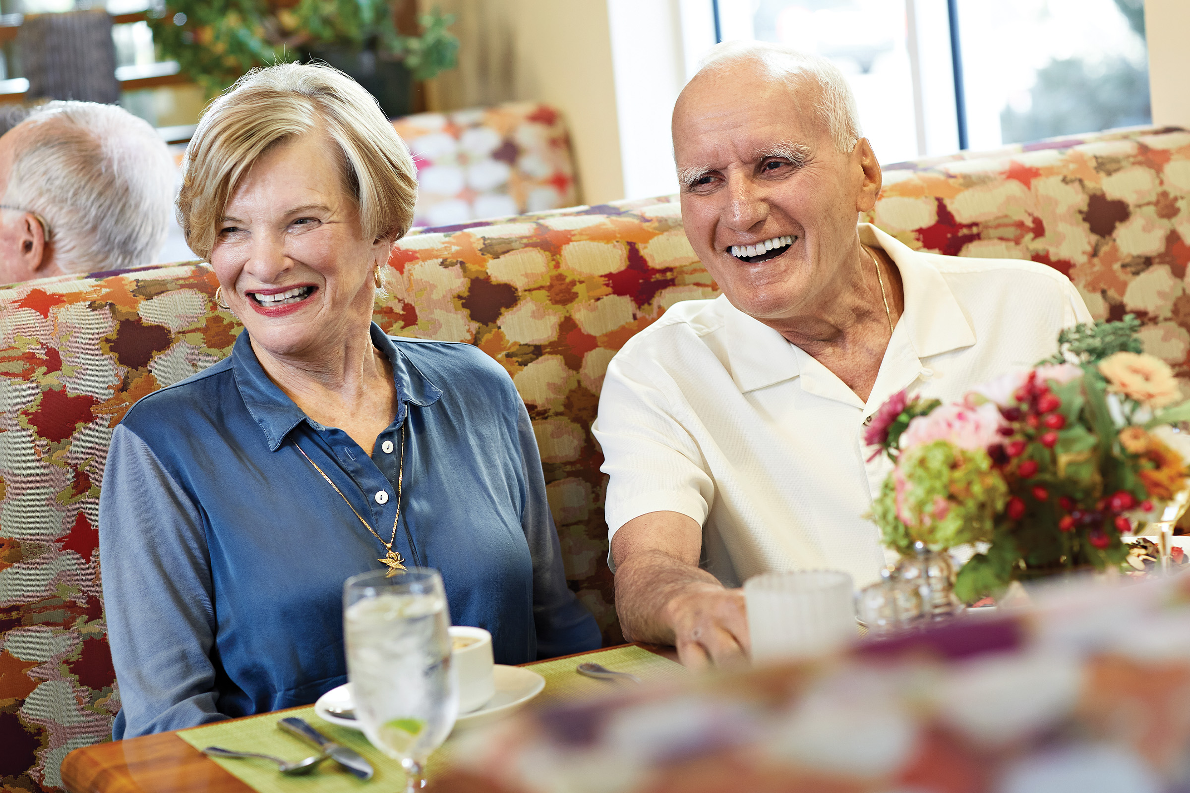 An older woman and man sit side by side in a restaurant booth, smiling and laughing. There are drinks, flowers, and dishes on the table. The atmosphere looks bright and cheerful.