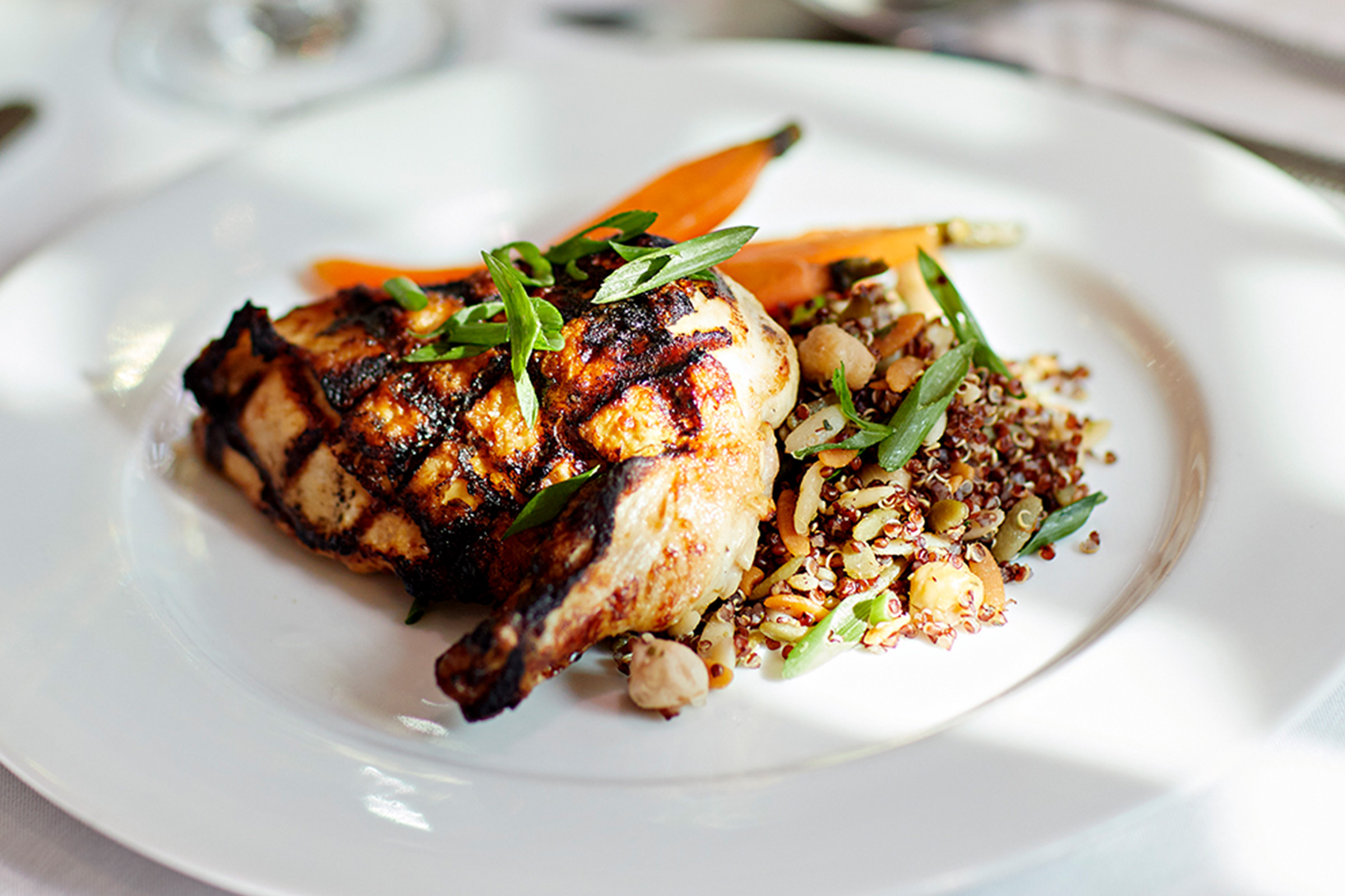 Grilled chicken with char marks, served with a side of quinoa salad, chopped nuts, green beans, and glazed carrots on a white plate. The dish is garnished with sliced green onions.