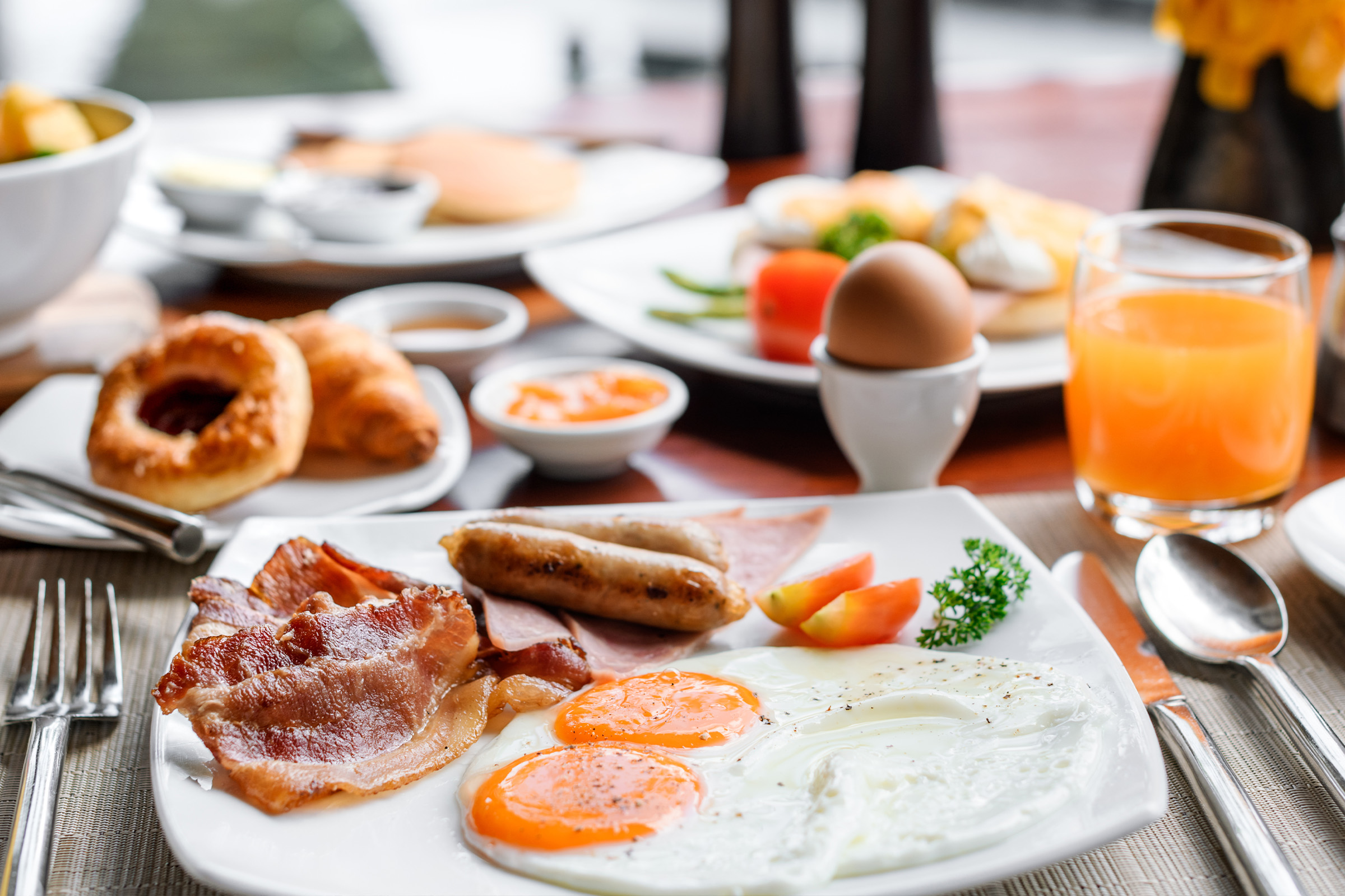 A breakfast plate with two sunny-side-up eggs, bacon, sausages, ham, a tomato slice, and parsley. An egg in an egg cup, croissant, orange juice, and condiments are also on the table.
