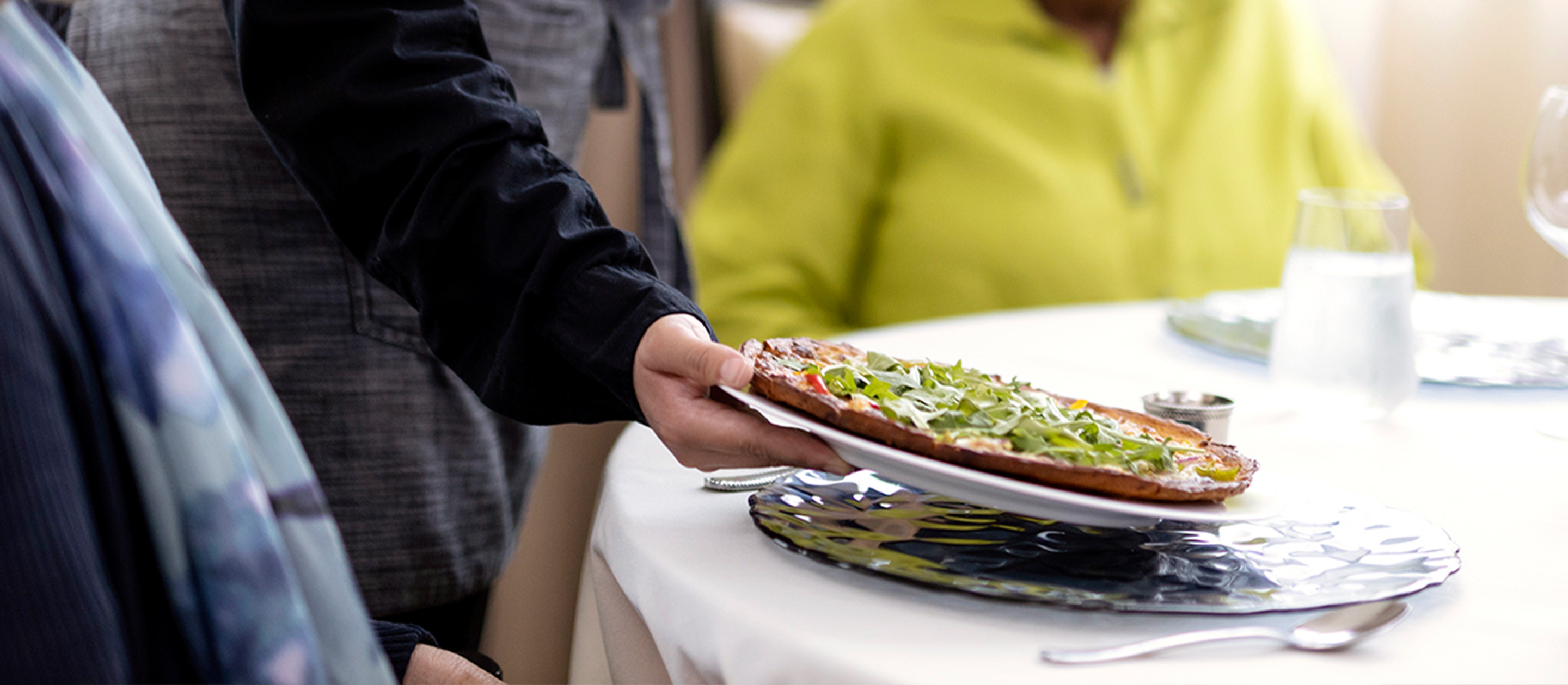 A person serves a plate with a flatbread topped with greens to a table where people are seated, one wearing a yellow top, with glassware and cutlery visible.