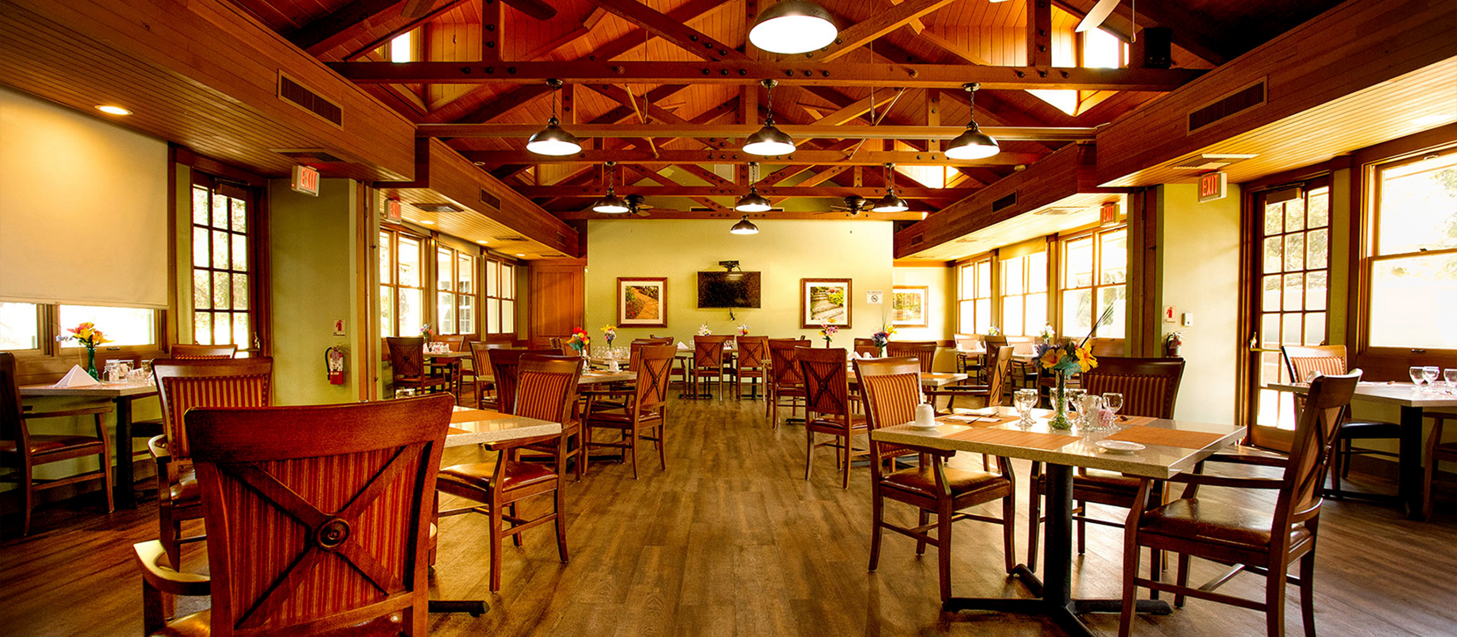 A spacious restaurant dining room with wooden floors, exposed beams, and large windows. Tables are set with glasses and napkins, and wooden chairs are neatly arranged. Warm natural light fills the inviting space.