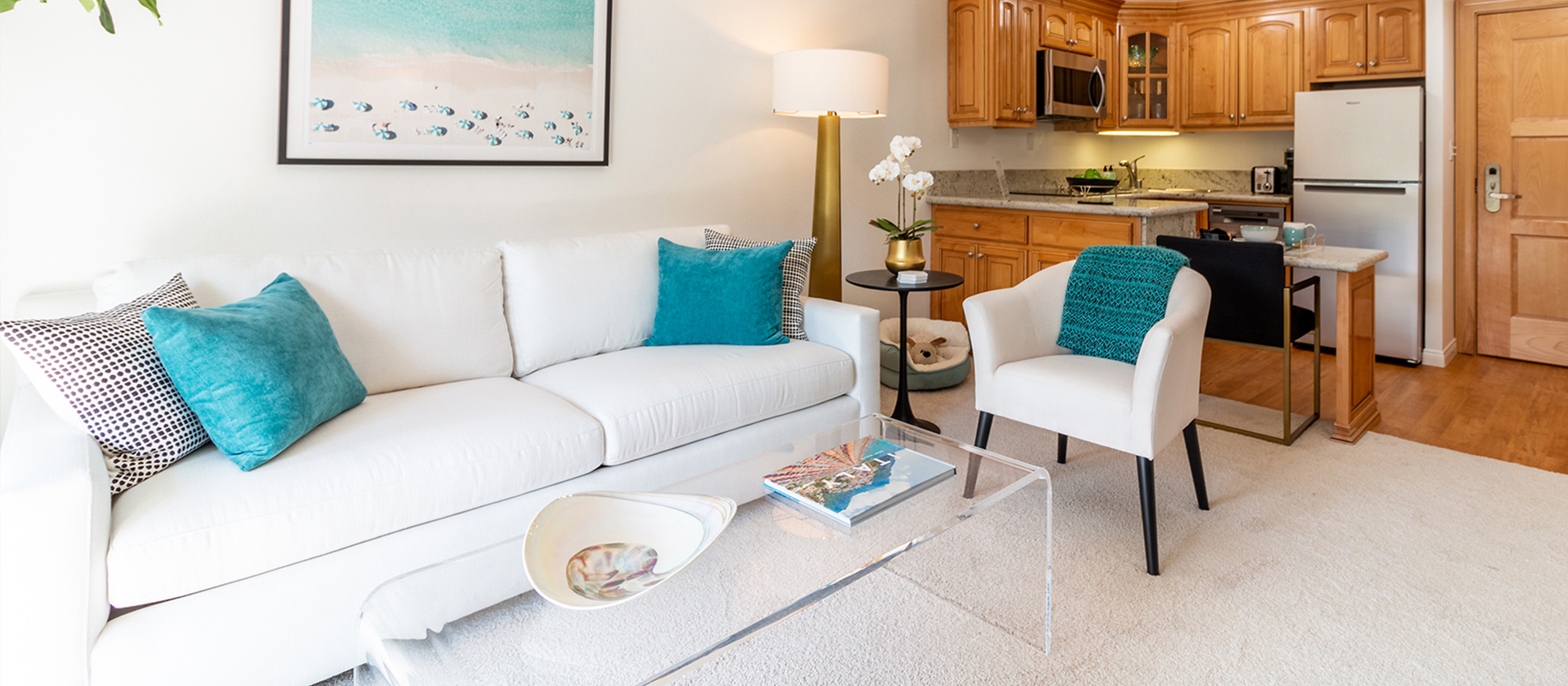 2.4_CAN_hero Bright living room with a white sofa, turquoise and black pillows, a modern armchair, clear coffee table, and decor. In the background, an open kitchen with wooden cabinets and stainless steel appliances.