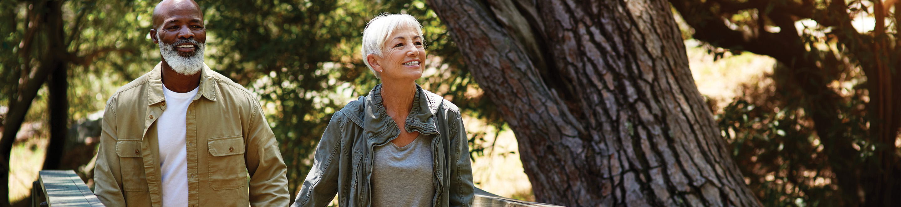An older couple walks outdoors on a sunny day, smiling and holding hands. They are surrounded by trees and greenery, enjoying a peaceful nature setting.