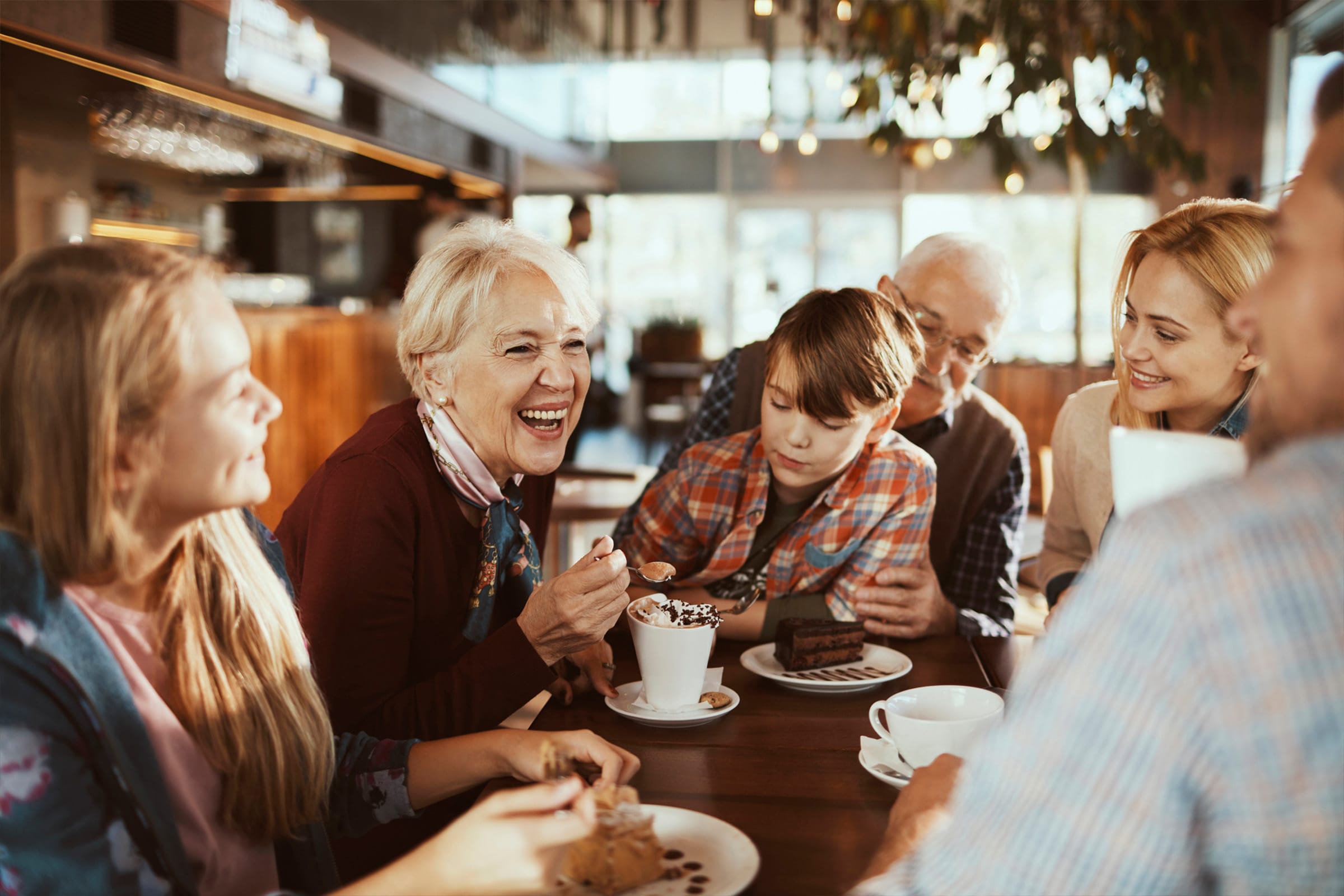 A group of people of different ages sit around a table in a cozy café, smiling and laughing while enjoying coffee, cake, and each other’s company. Sunlight streams through the windows in the background.