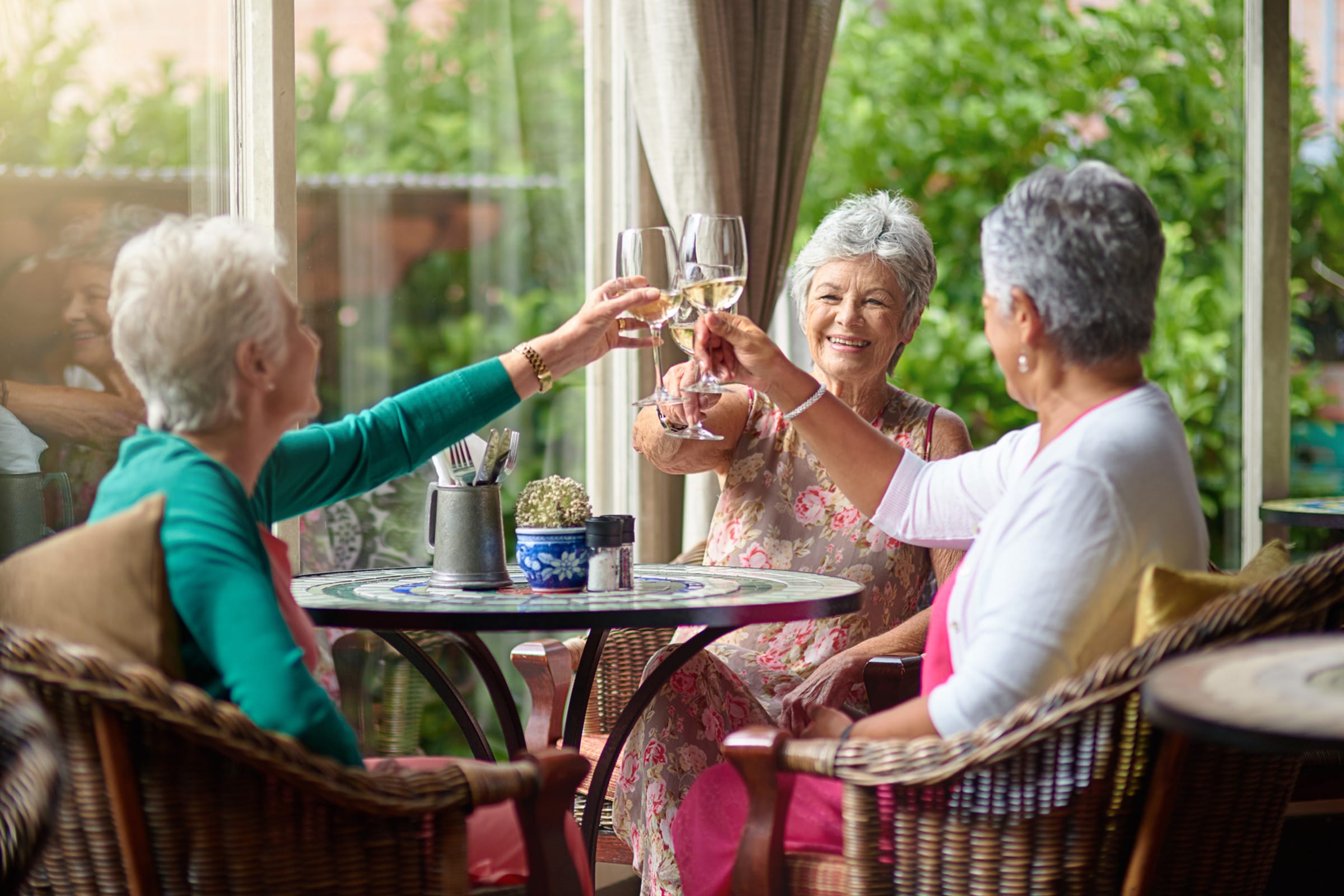 2.6_CO_photo Three older women sit around a table at an outdoor cafe, smiling and raising glasses in a toast. Sunlight streams in, and green plants are visible in the background.