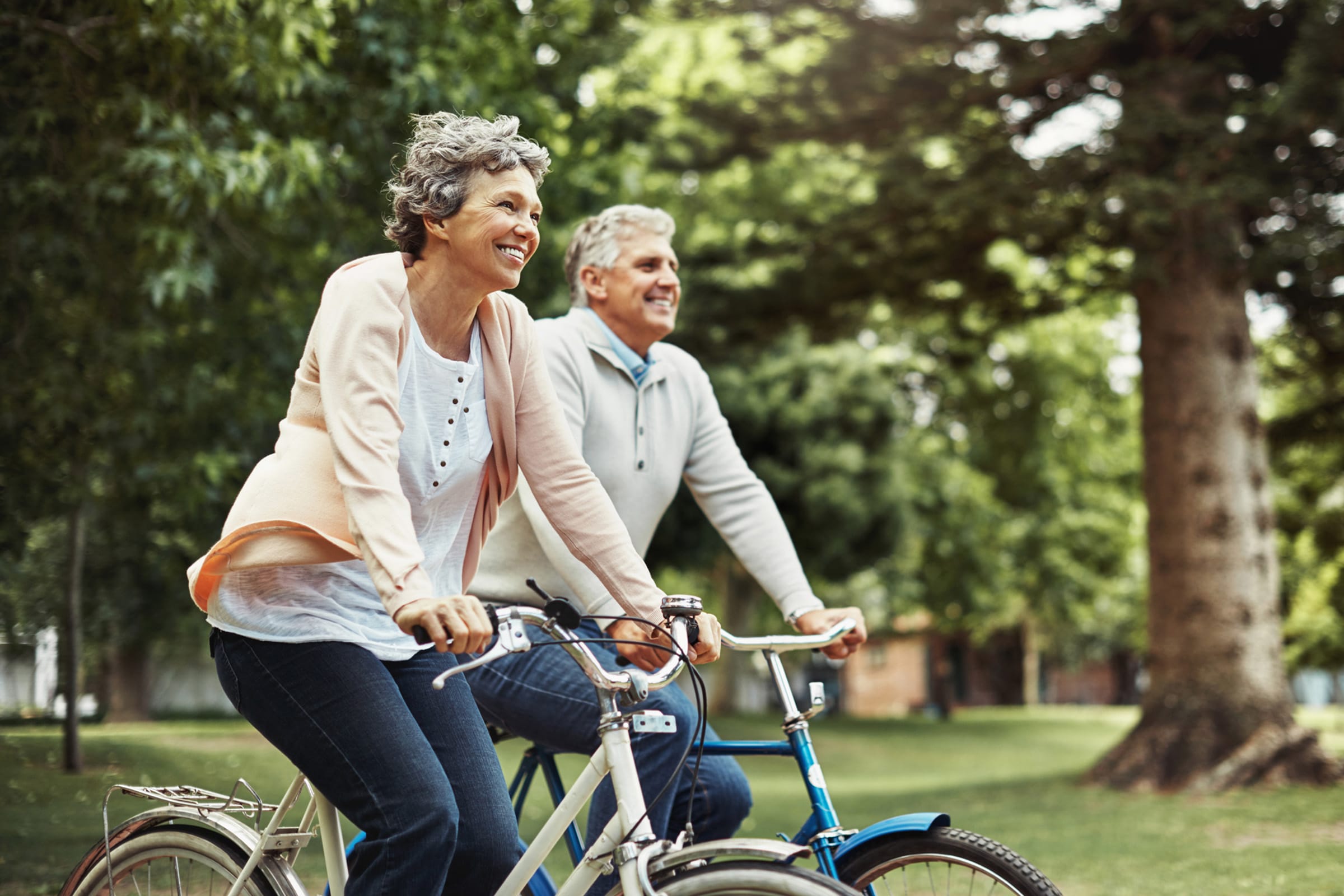 Two older adults ride bicycles through a green park, smiling and enjoying the outdoors. Trees and grass surround them, creating a peaceful and lively atmosphere.