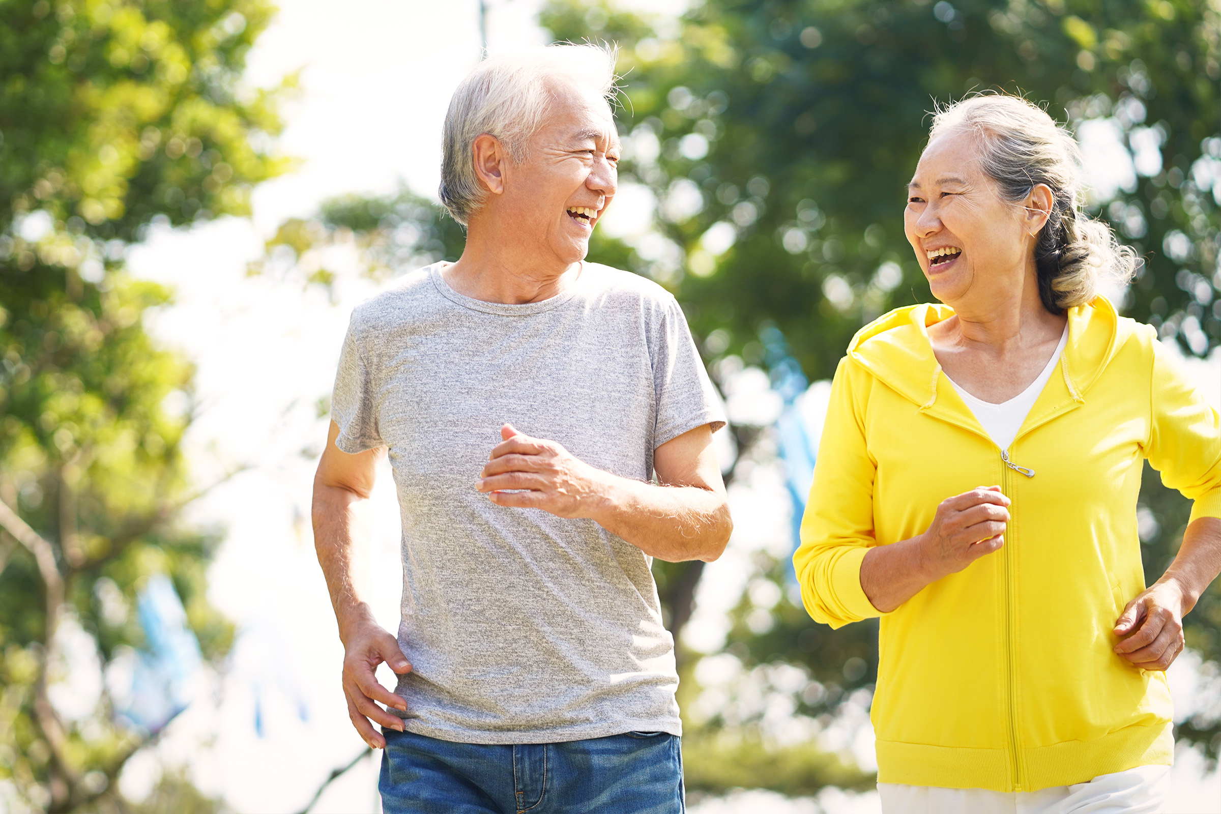 An older man and woman are smiling and jogging outdoors on a sunny day, surrounded by green trees. Both appear happy and energetic, enjoying their time together in the park.