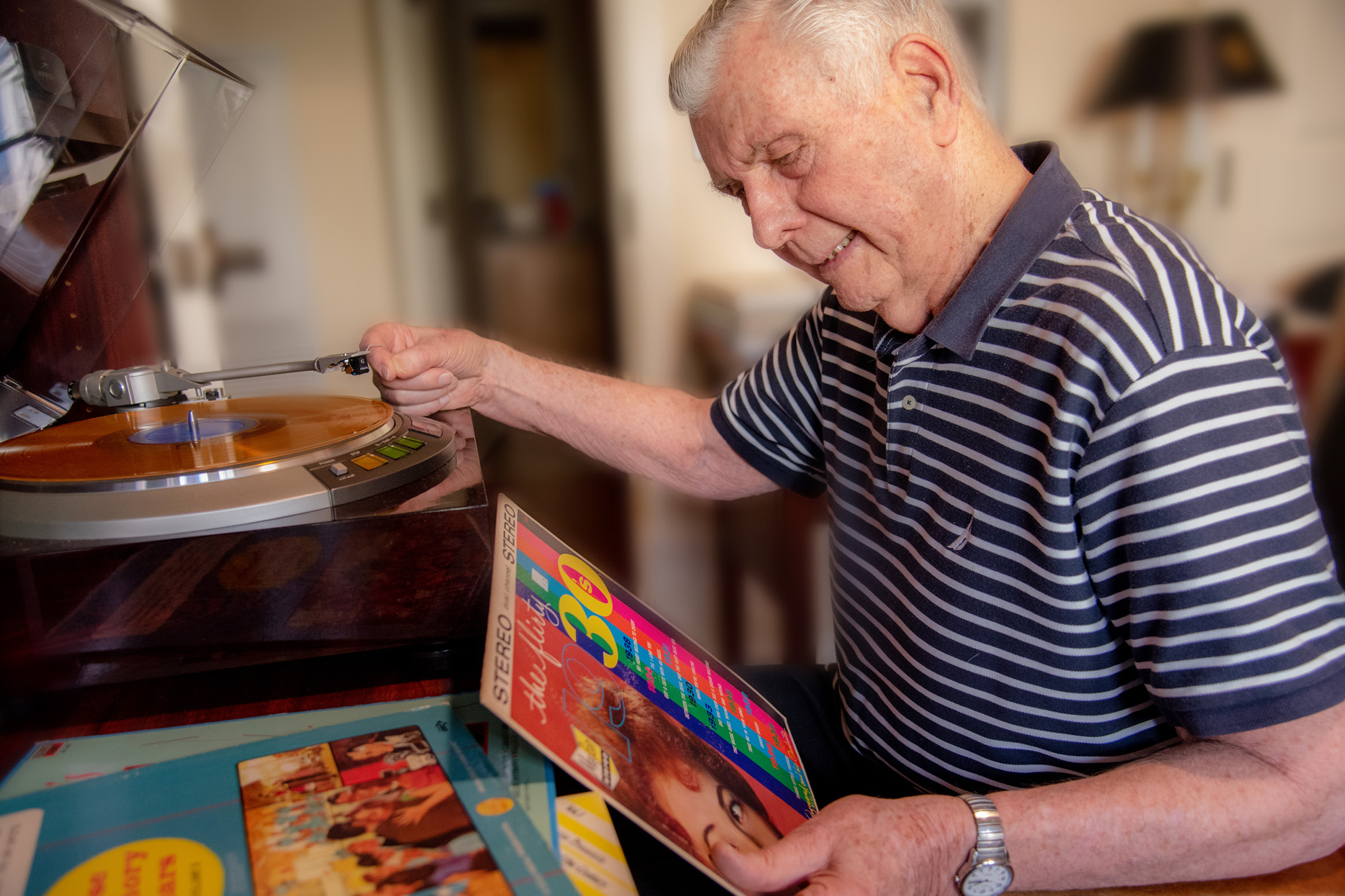 An elderly man in a striped polo shirt smiles as he plays a vinyl record on a turntable, holding a colorful record sleeve, with more vinyl albums stacked nearby.