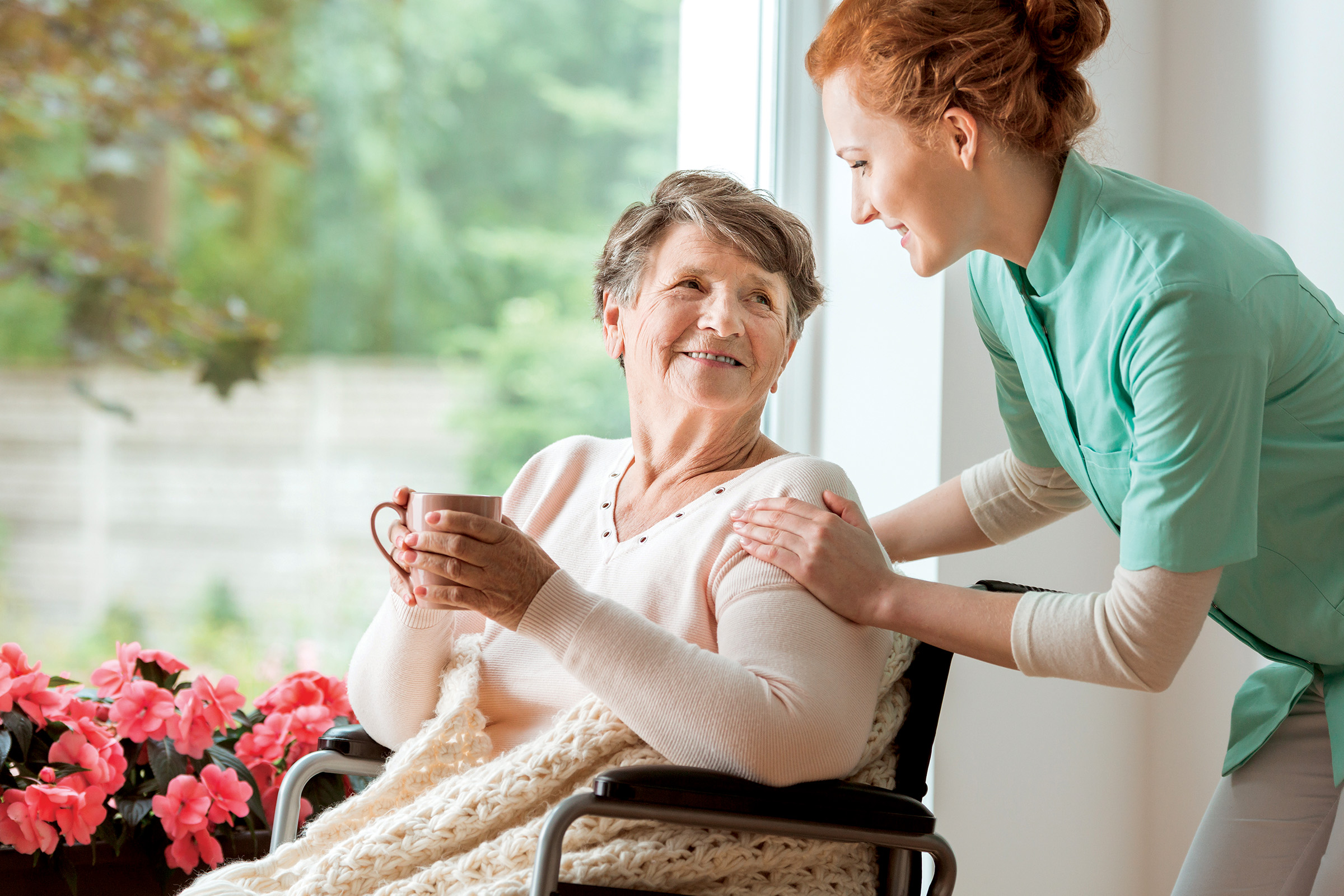 A smiling elderly woman sits in a wheelchair holding a mug, covered with a knit blanket. A caregiver in a green uniform stands beside her, smiling and touching her shoulder. Pink flowers are on the windowsill nearby.