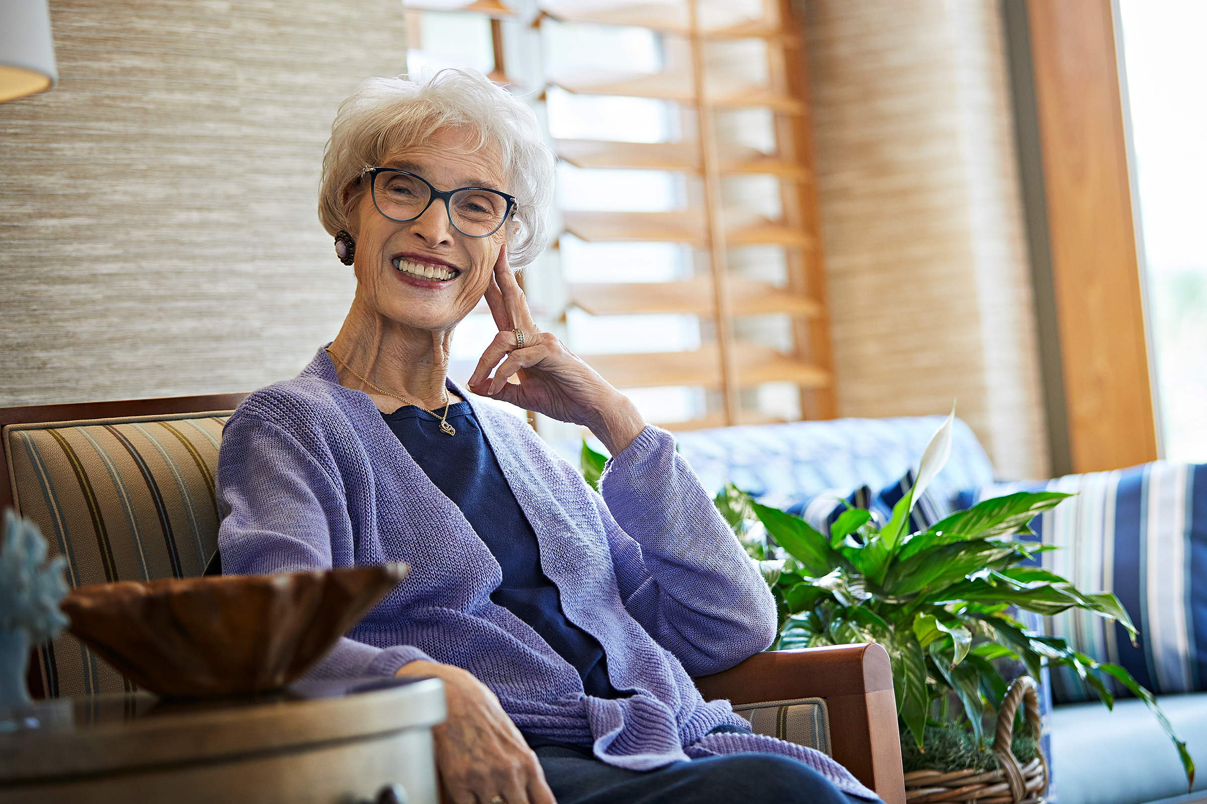 Elderly woman with short gray hair and glasses, smiling and sitting on a striped chair indoors, wearing a purple cardigan. Green potted plant and natural light fill the cozy, well-lit room.