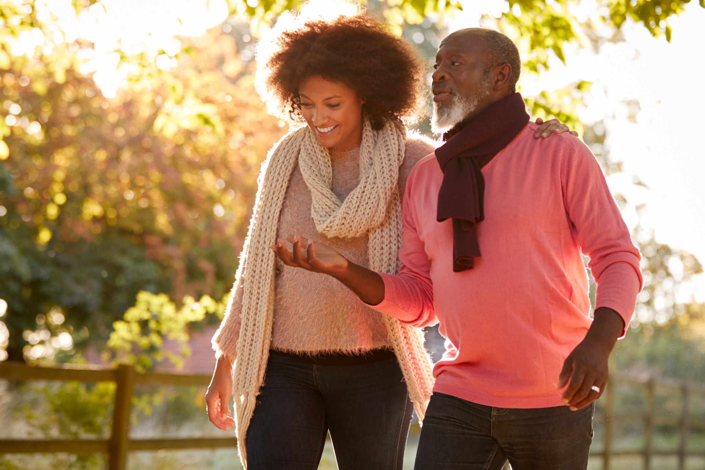 A smiling woman and man walk outdoors together on a sunny day, dressed warmly in sweaters and scarves, enjoying each others company in a park-like setting with autumn foliage in the background.