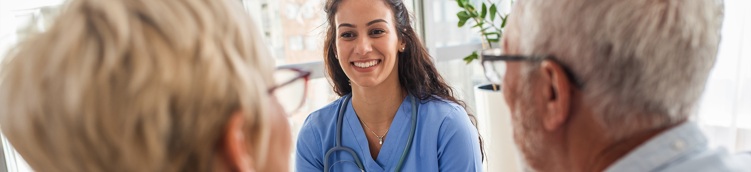 A smiling healthcare professional in blue scrubs talks with two older adults in a bright, modern setting. The professional has a stethoscope around her neck.