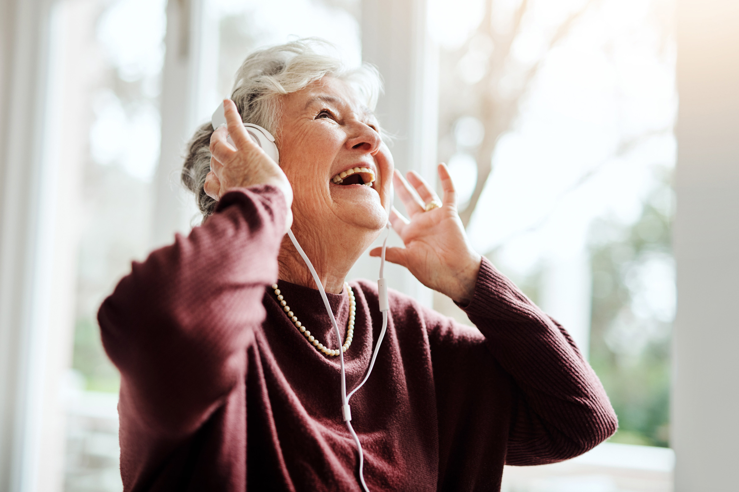An elderly woman with gray hair, wearing a maroon sweater and pearl necklace, smiles joyfully while listening to music on headphones in a bright, sunlit room.