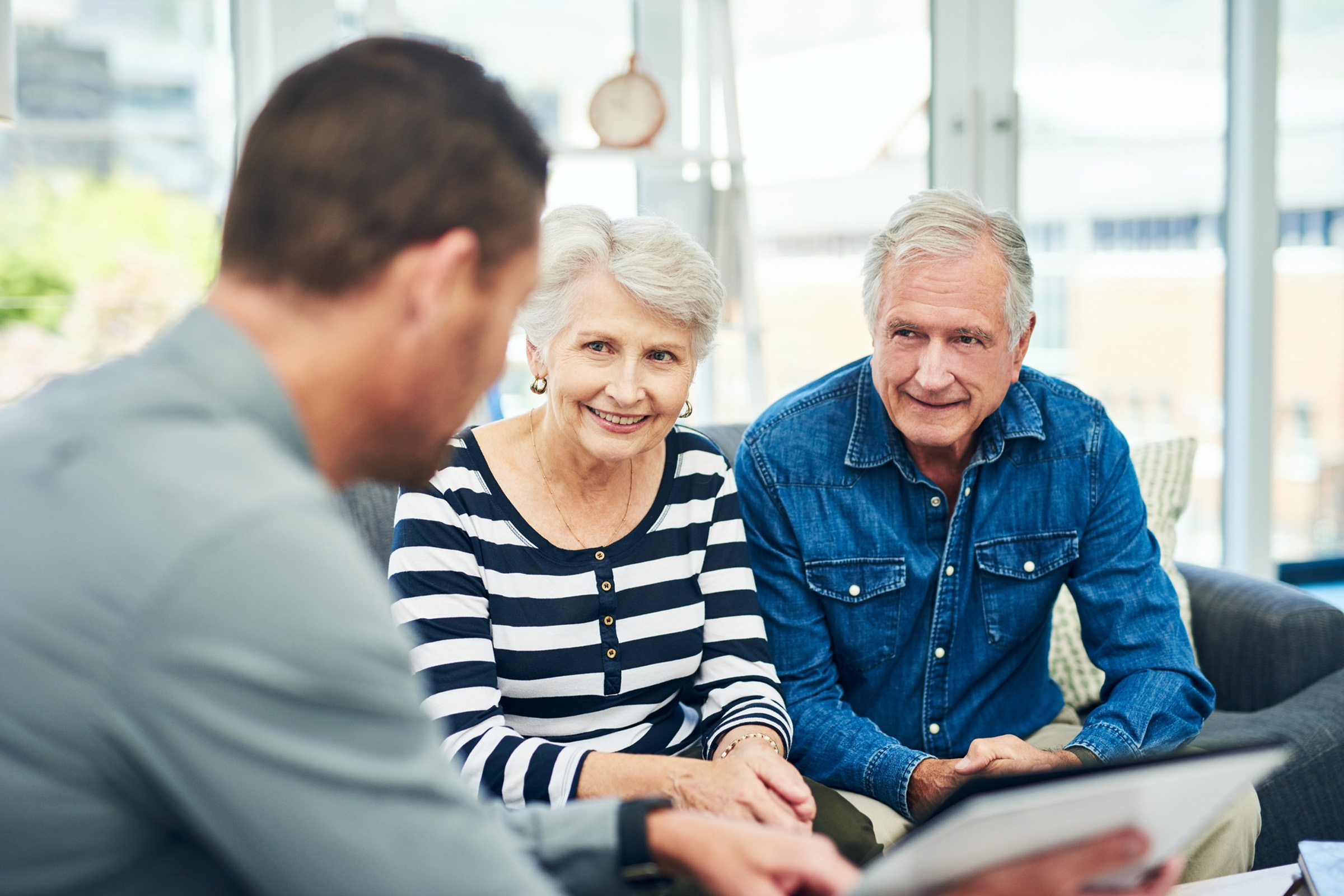 An older couple sits on a couch, smiling and listening to a man holding a tablet, possibly discussing finances or plans in a bright, modern living room.