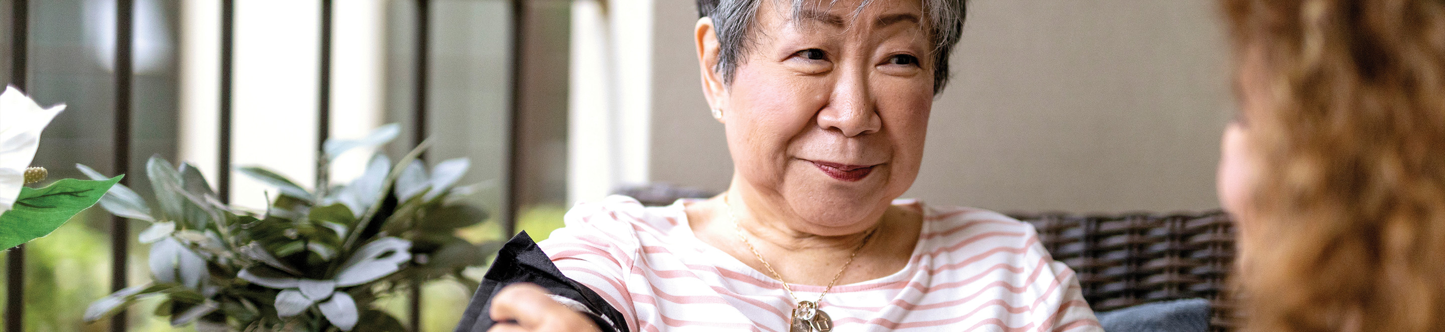 An older woman with short gray hair and a striped shirt sits on a patio, smiling at a person with curly hair. There are green plants and a wicker chair in the background.