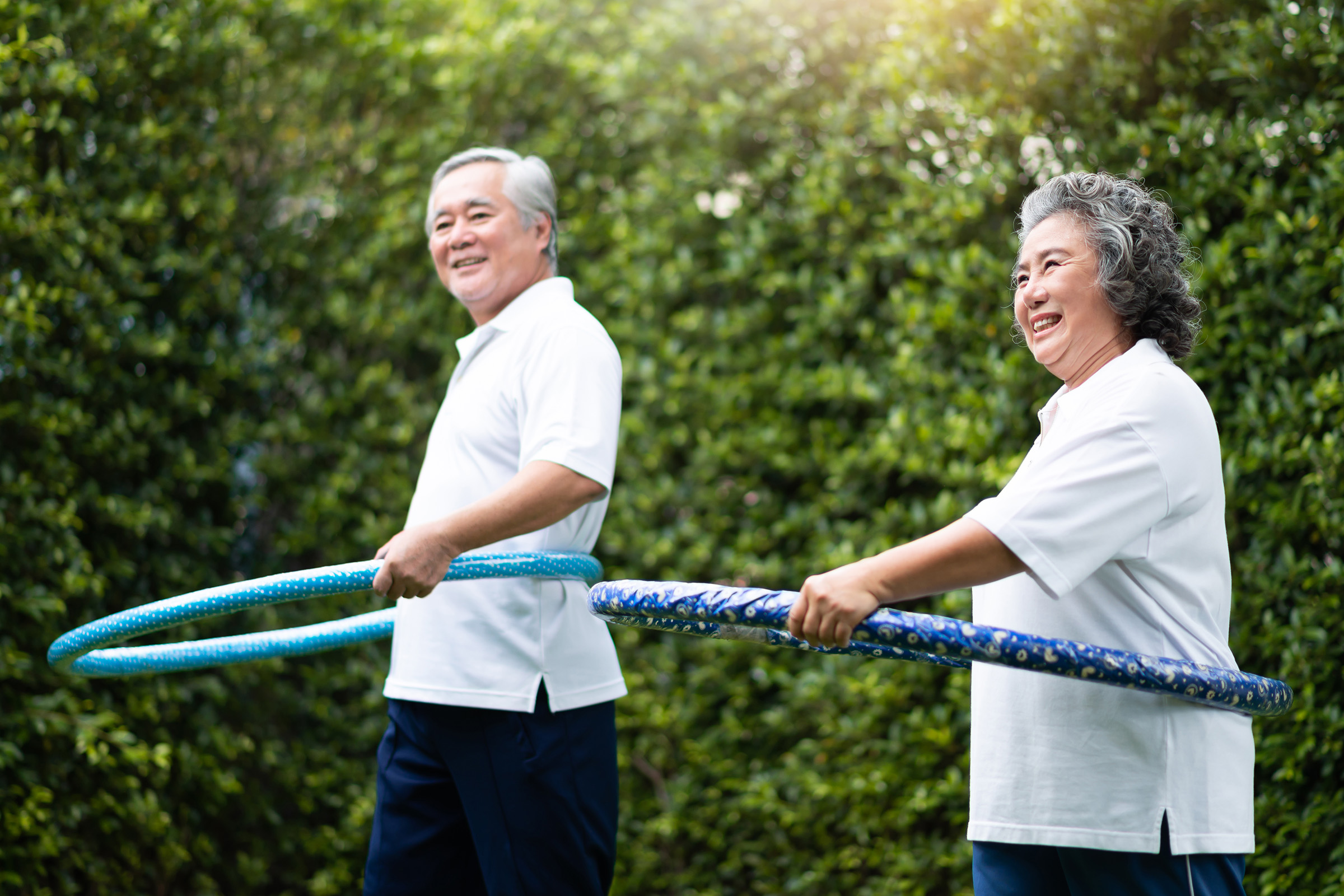 Two smiling older adults in white shirts and dark pants use hula hoops outdoors, standing side by side in front of green foliage on a sunny day.