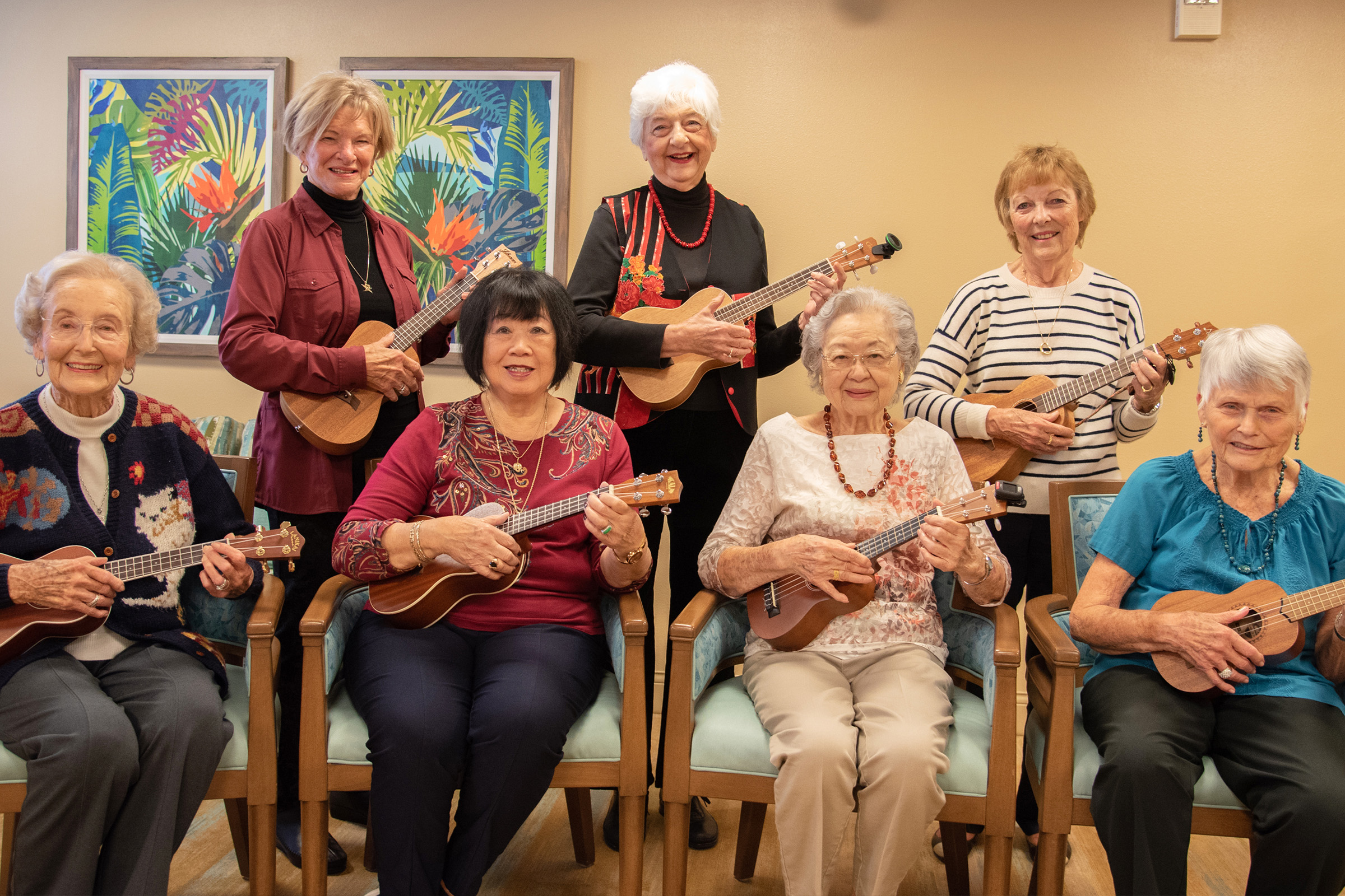 Seven elderly women, smiling and seated or standing, play ukuleles in a cozy room with colorful botanical artwork on the wall behind them. They appear joyful and relaxed, wearing casual, colorful clothing.