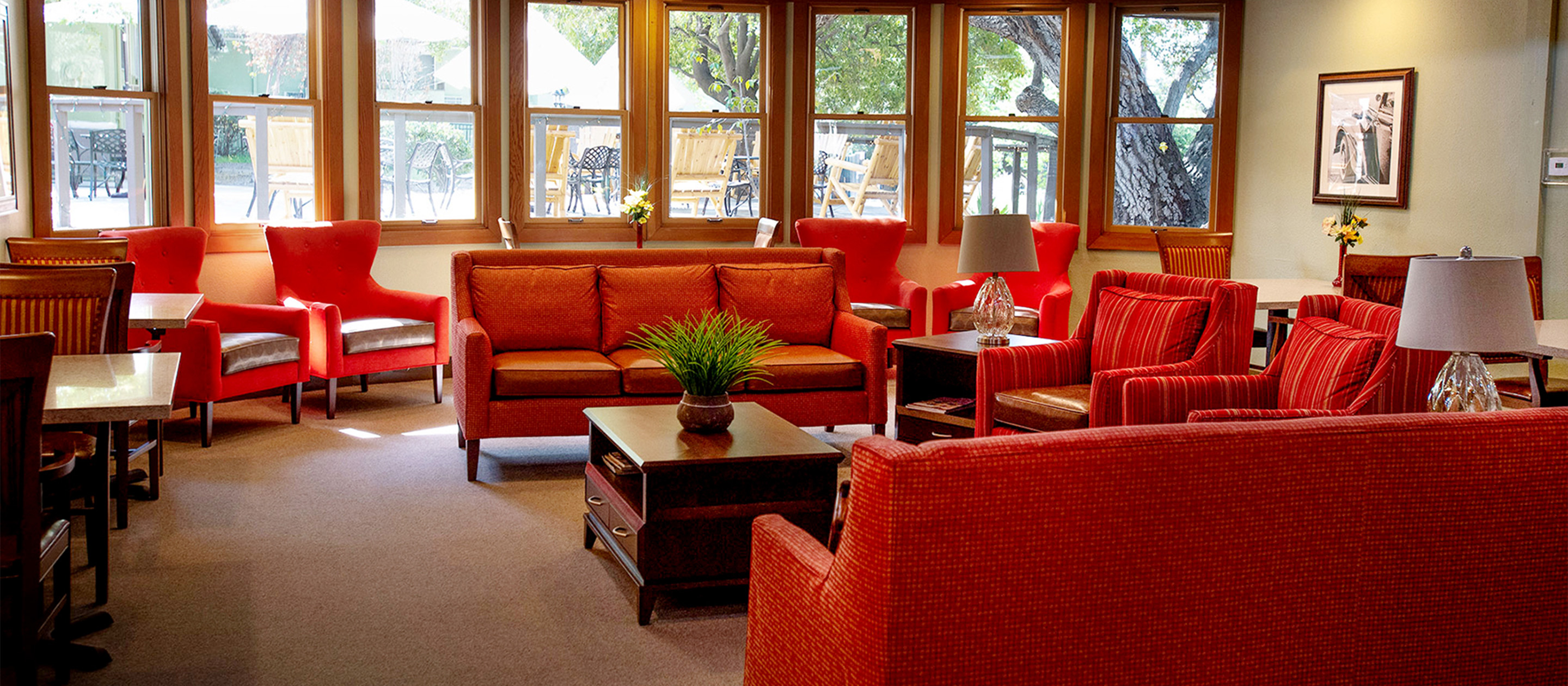 A cozy lounge area with red sofas and armchairs, wooden tables, two lamps, and a potted plant on a coffee table. Large windows let in natural light and overlook trees and an outdoor patio with tables and chairs.