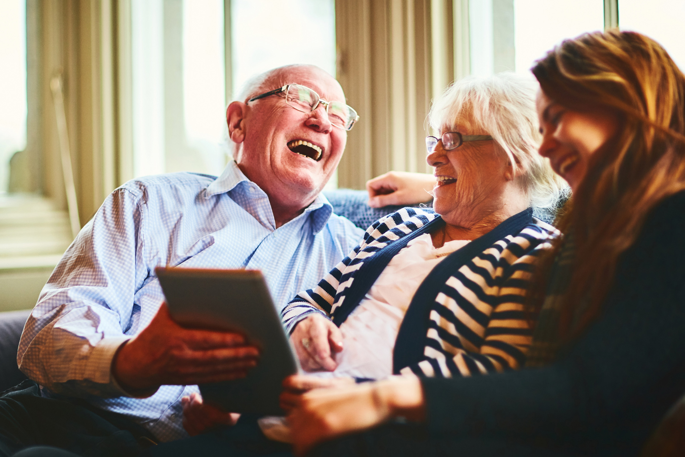 An elderly man and woman sit on a couch with a younger woman, all laughing together while looking at a tablet. Sunlight streams through large windows behind them, creating a warm and joyful atmosphere.