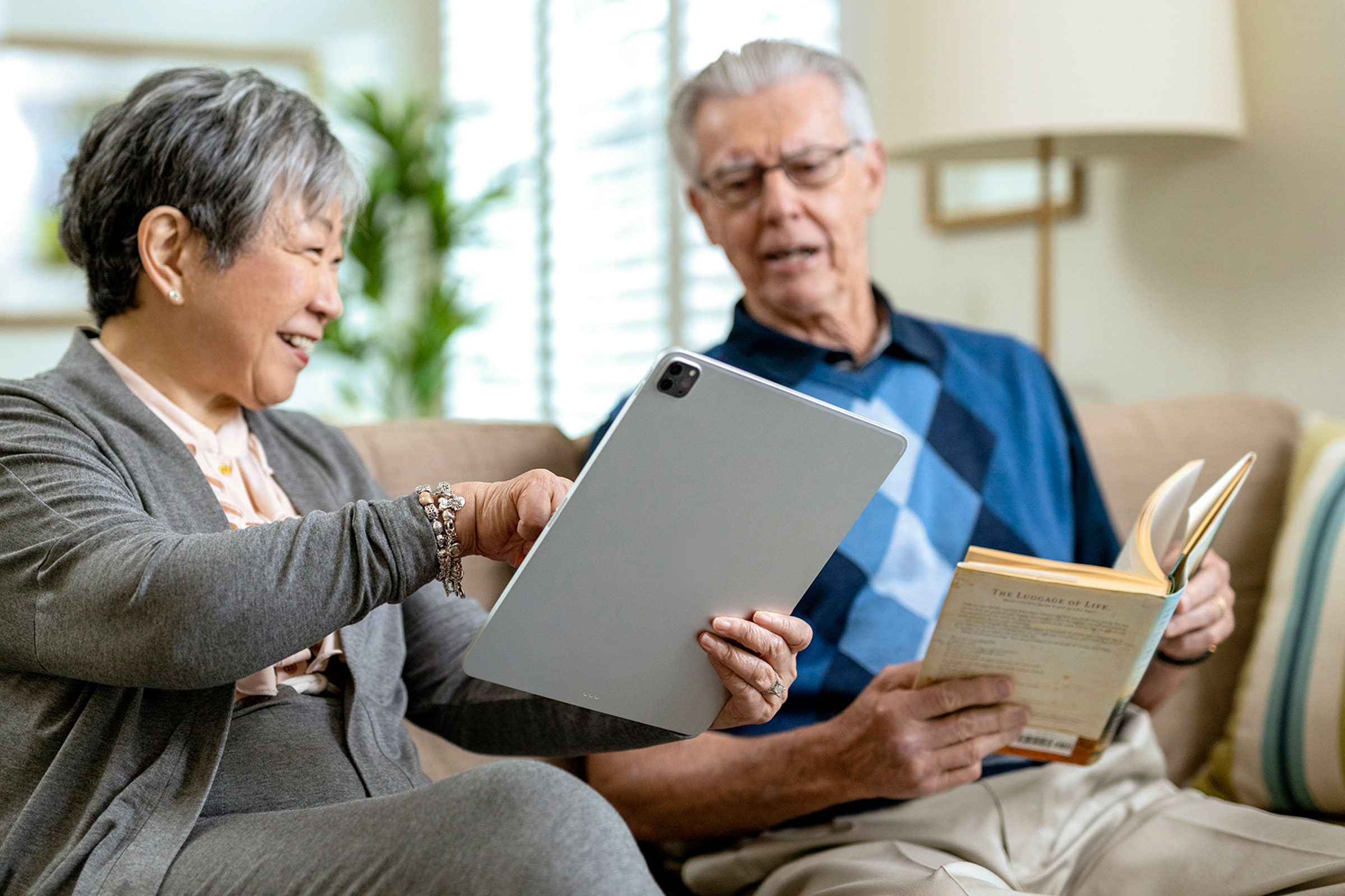 An older woman and man sit together on a couch. The woman is smiling and pointing at a tablet, while the man holds an open book, looking at the screen. They appear relaxed and engaged in a living room setting.