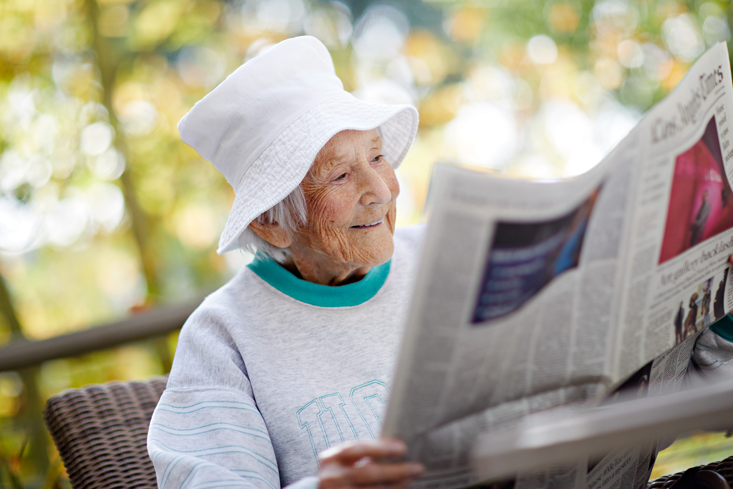 An elderly woman in a white hat and grey sweatshirt sits outdoors, smiling as she reads a newspaper. The background is blurred with greenery and sunlight.