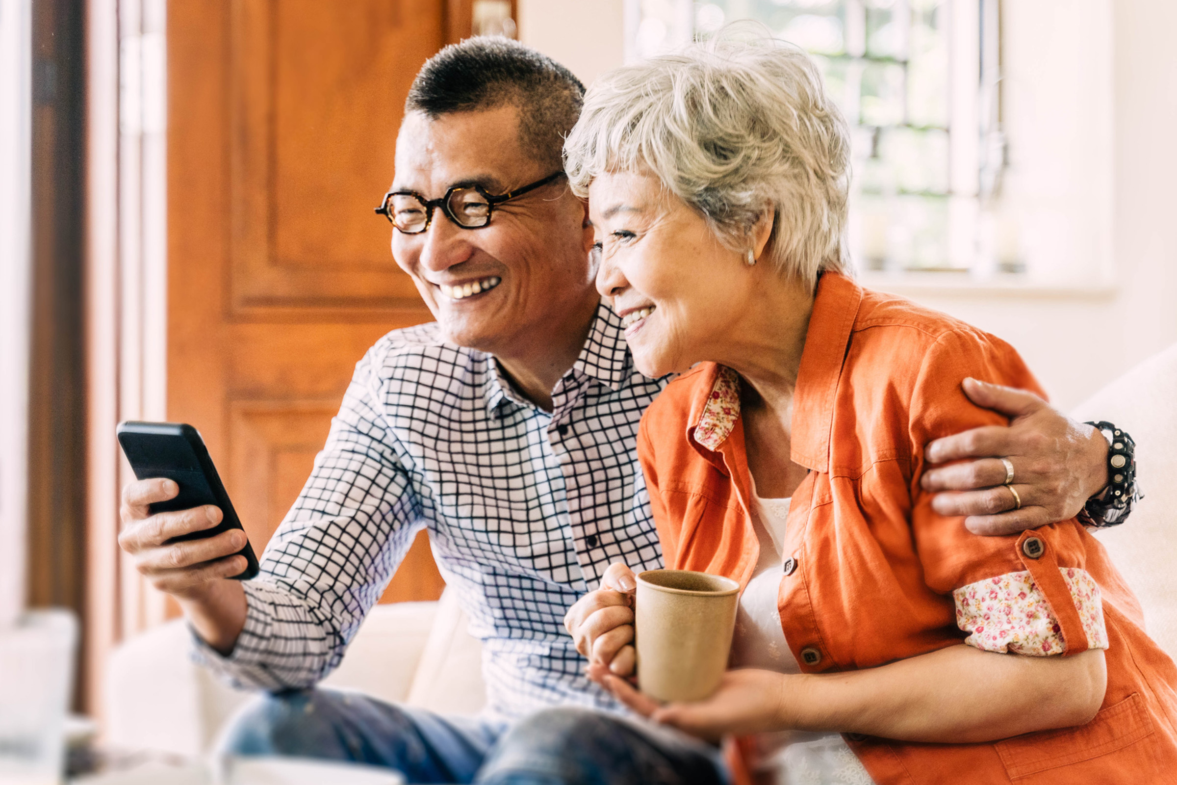 An older man and woman sit together on a couch, smiling as they look at a smartphone. The woman holds a mug while the man wraps his arm around her. Sunlight streams through a window behind them.