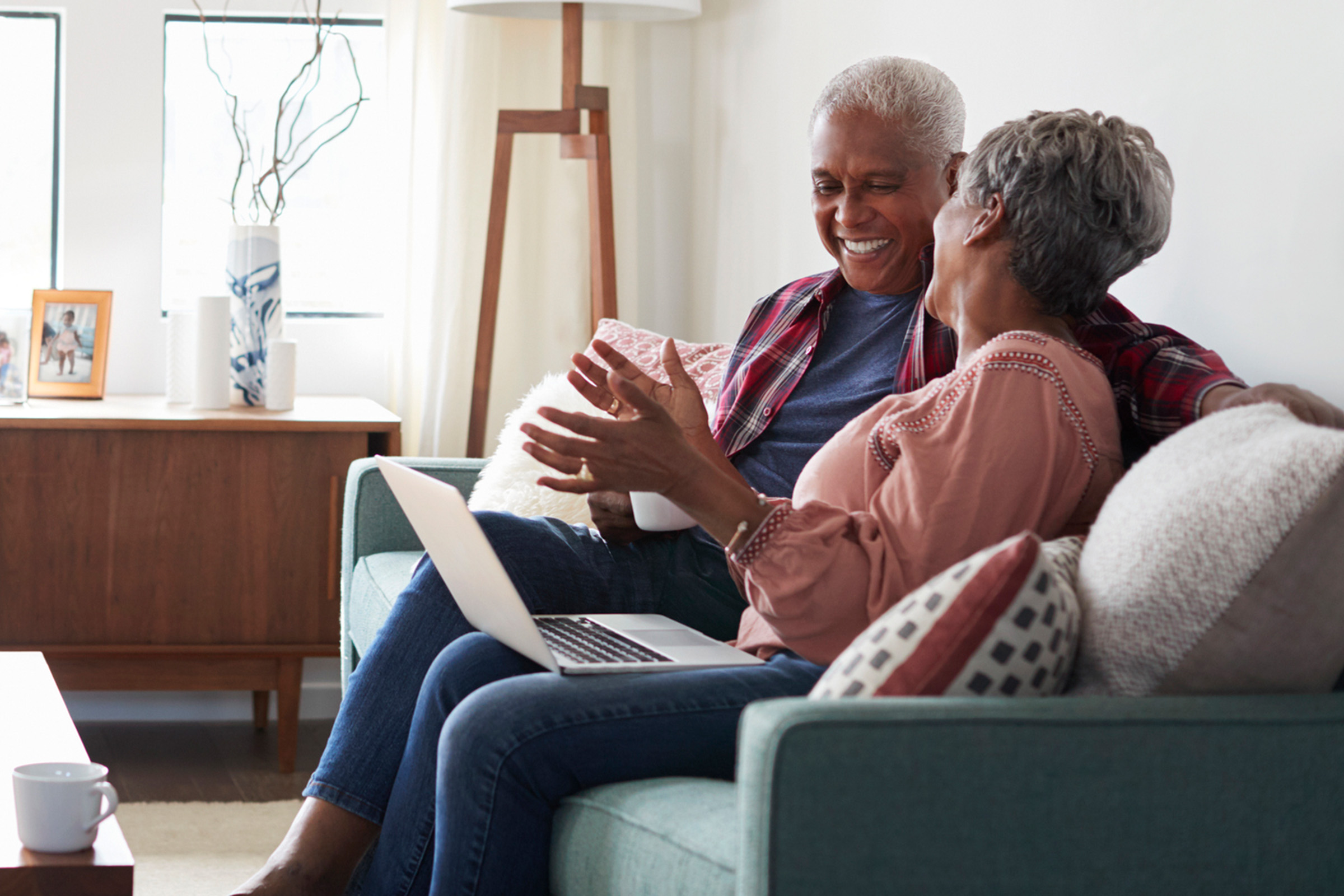 An older couple sits on a couch at home, smiling and talking while looking at a laptop. One holds a coffee mug, and the room is cozy with natural light and modern decor.