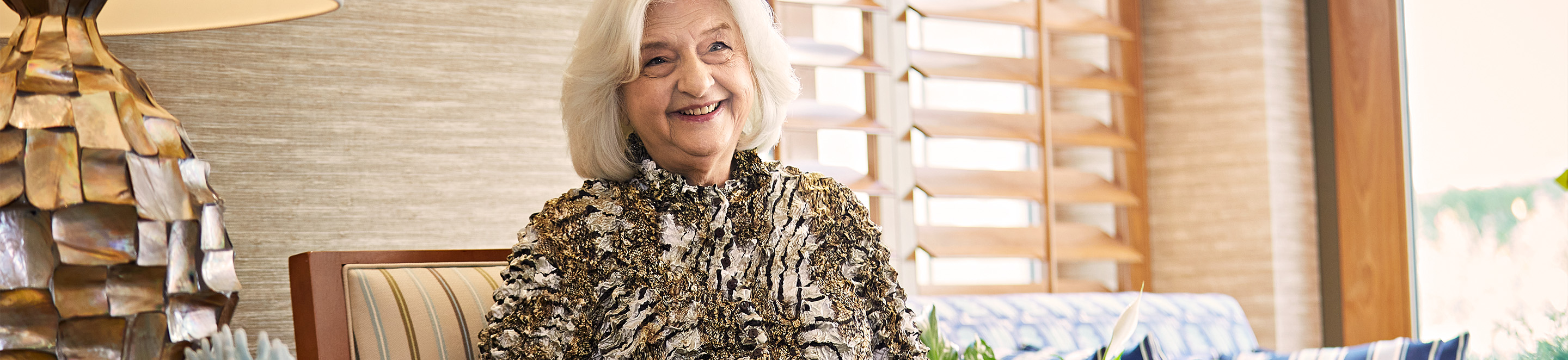 An older woman with white hair, wearing a patterned top, sits and smiles on a striped chair indoors near a window with wooden blinds and a decorative lamp.