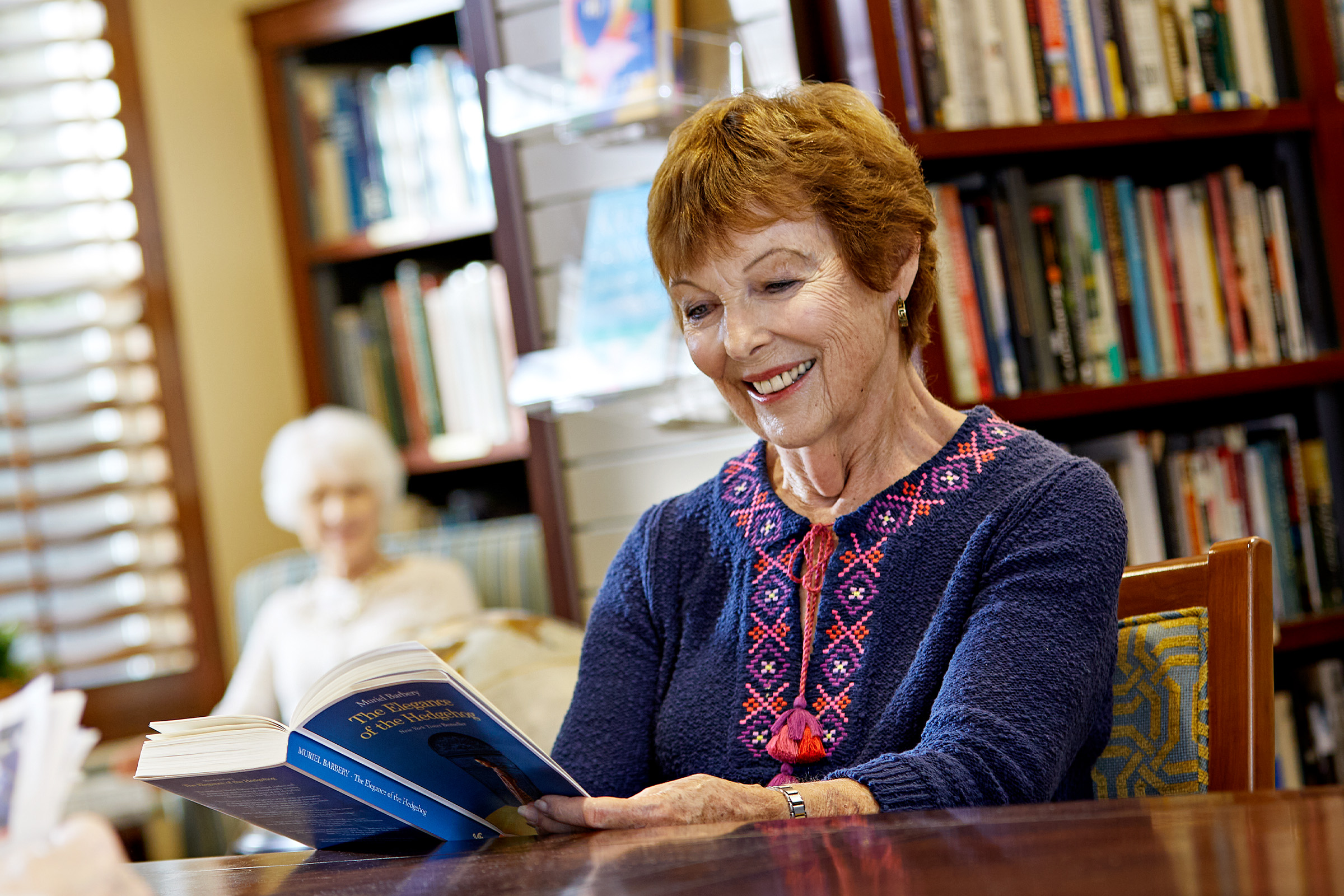 An older woman with short brown hair, wearing a blue sweater with a pink pattern, sits at a table smiling while reading a book. Bookshelves and another elderly woman are visible in the background.