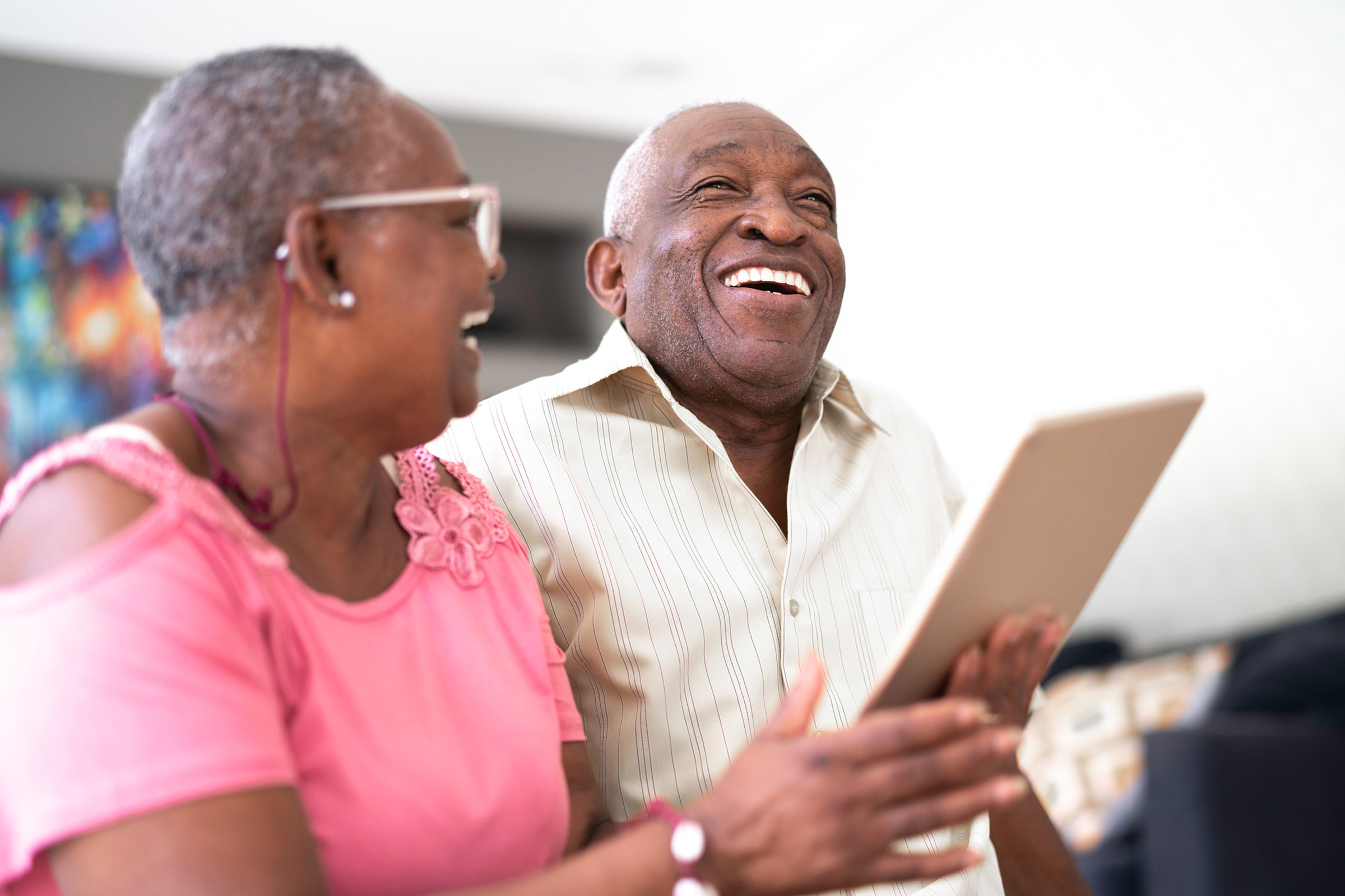 An older couple sits together indoors, smiling and laughing while holding a tablet. The woman wears a pink top and glasses, while the man wears a light striped shirt. They appear to be enjoying a joyful moment together.