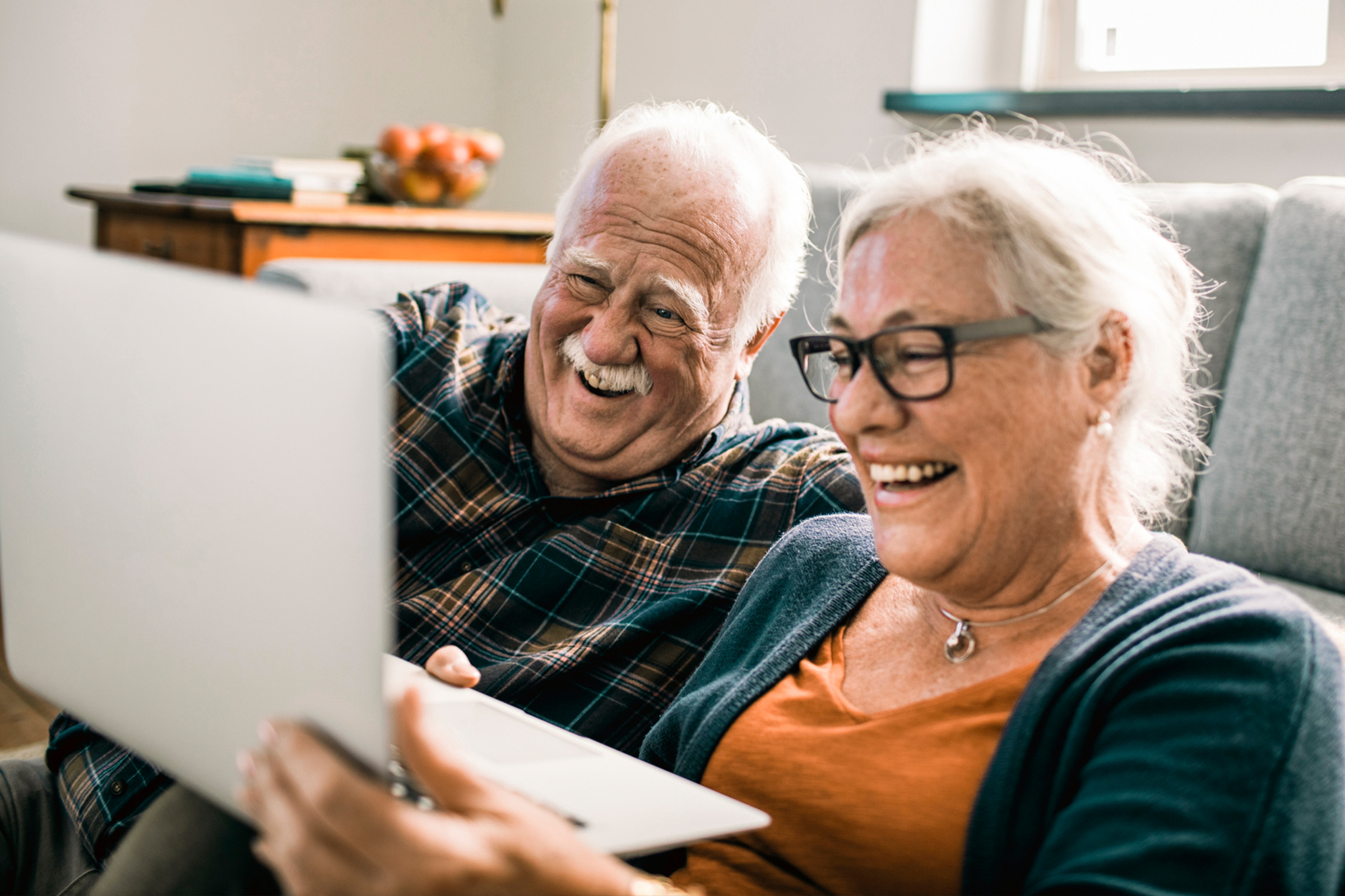 Two smiling older adults sit close together on a couch, looking at a laptop screen and laughing. The room appears cozy with warm lighting and a bowl of fruit in the background.