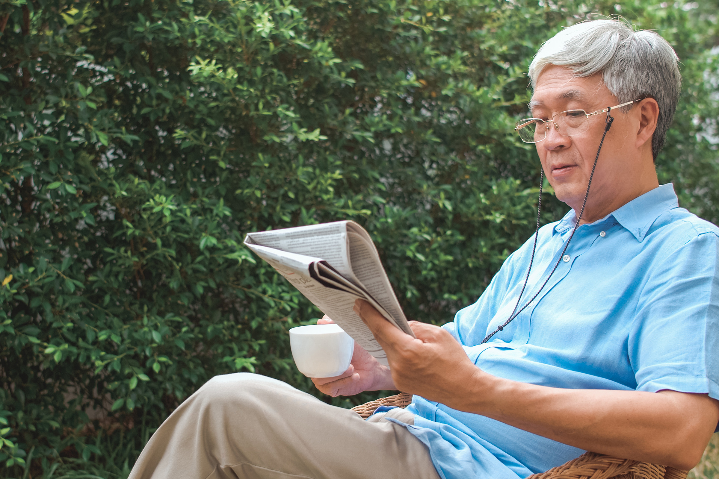 An older man with gray hair and glasses sits outdoors on a wicker chair, reading a newspaper and holding a white mug. He is wearing a light blue shirt and appears relaxed, surrounded by green foliage.