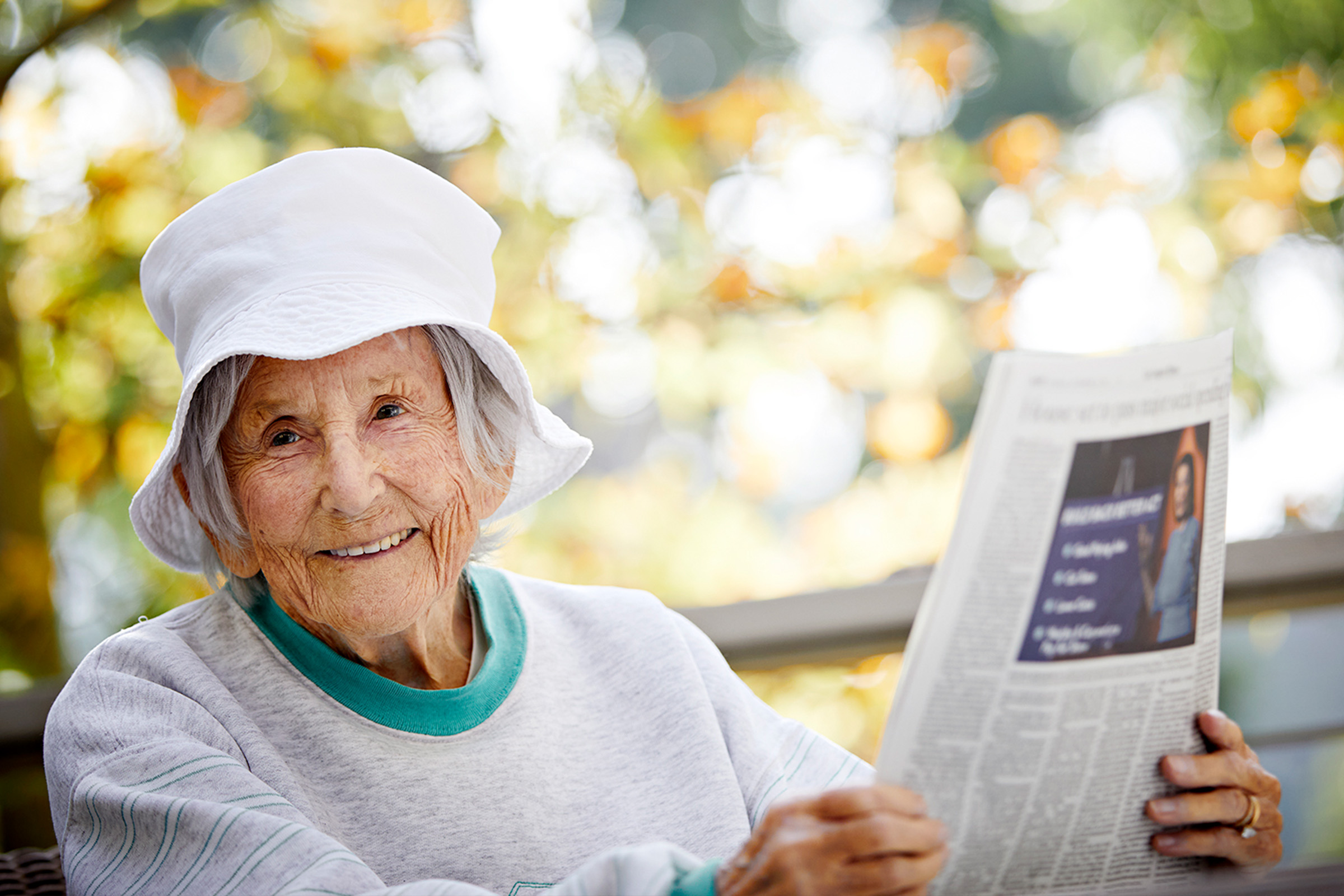 An elderly woman wearing a white hat and light sweater smiles while sitting outdoors, holding and reading a newspaper. The background is softly blurred with trees and sunlight.