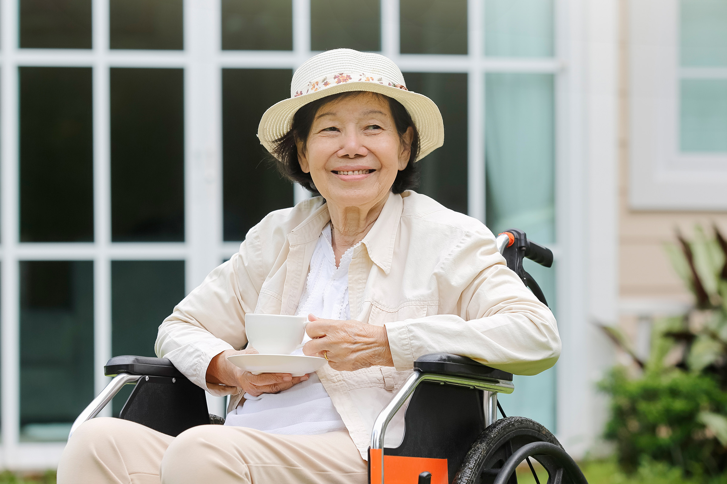 An older woman in a hat smiles while sitting in a wheelchair outdoors, holding a cup and saucer, with a house and greenery visible in the background.