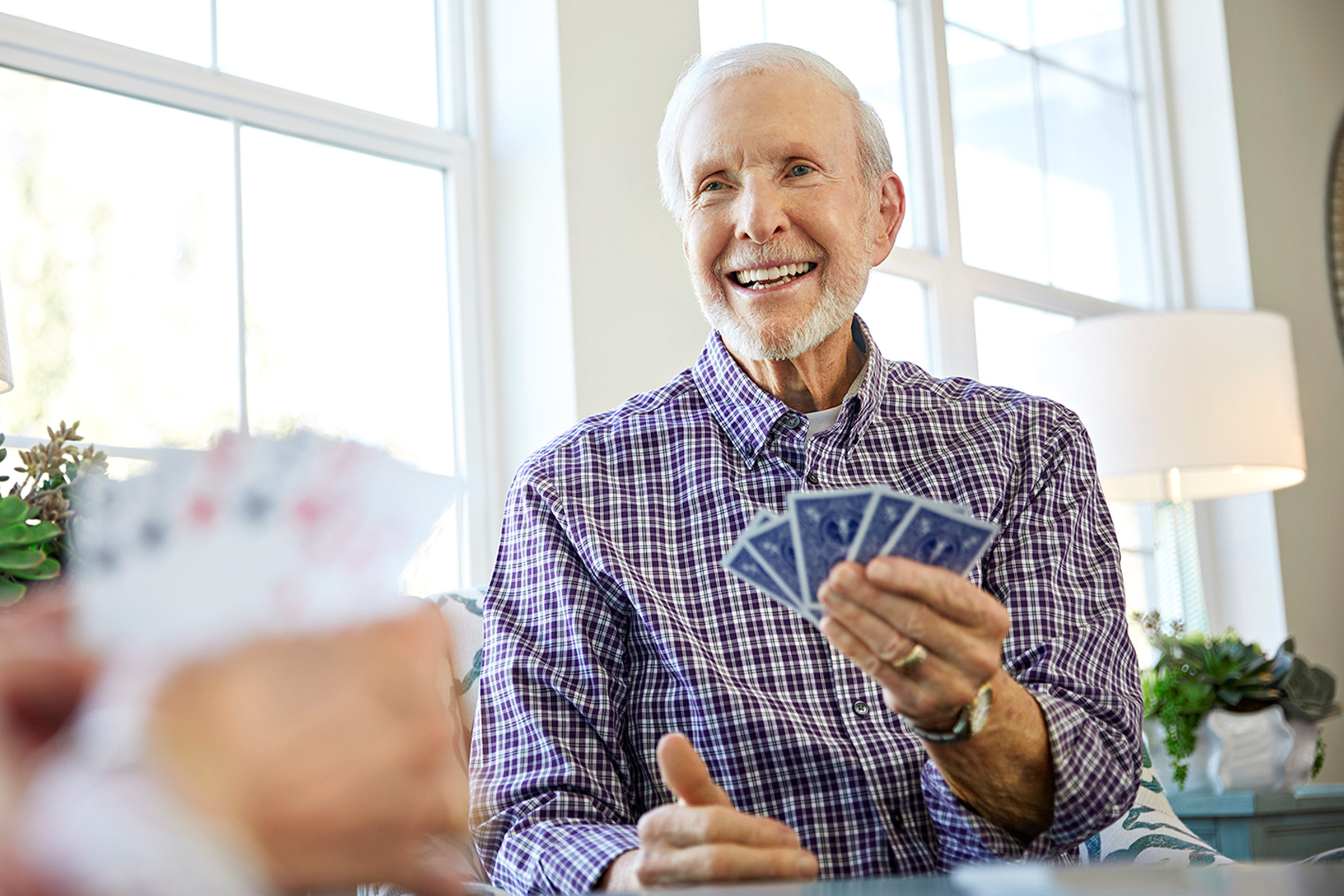 An older man in a plaid shirt smiles while holding playing cards, seated at a table with another player. Sunlight streams in through large windows behind him, creating a bright and cheerful atmosphere.