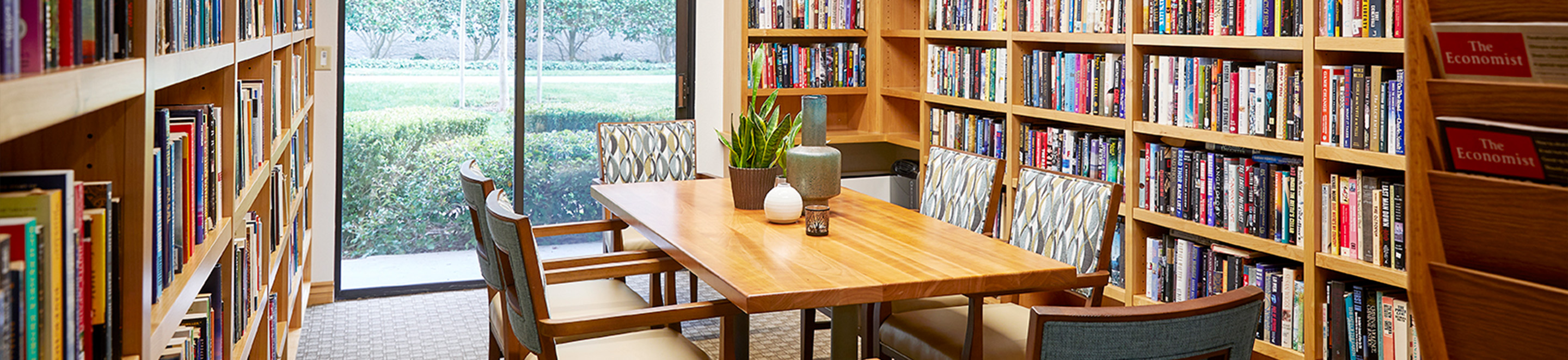 A cozy library room with wooden bookshelves full of books, a rectangular wooden table, four cushioned chairs, and a large window overlooking green bushes outside. A vase with a plant sits on the table.