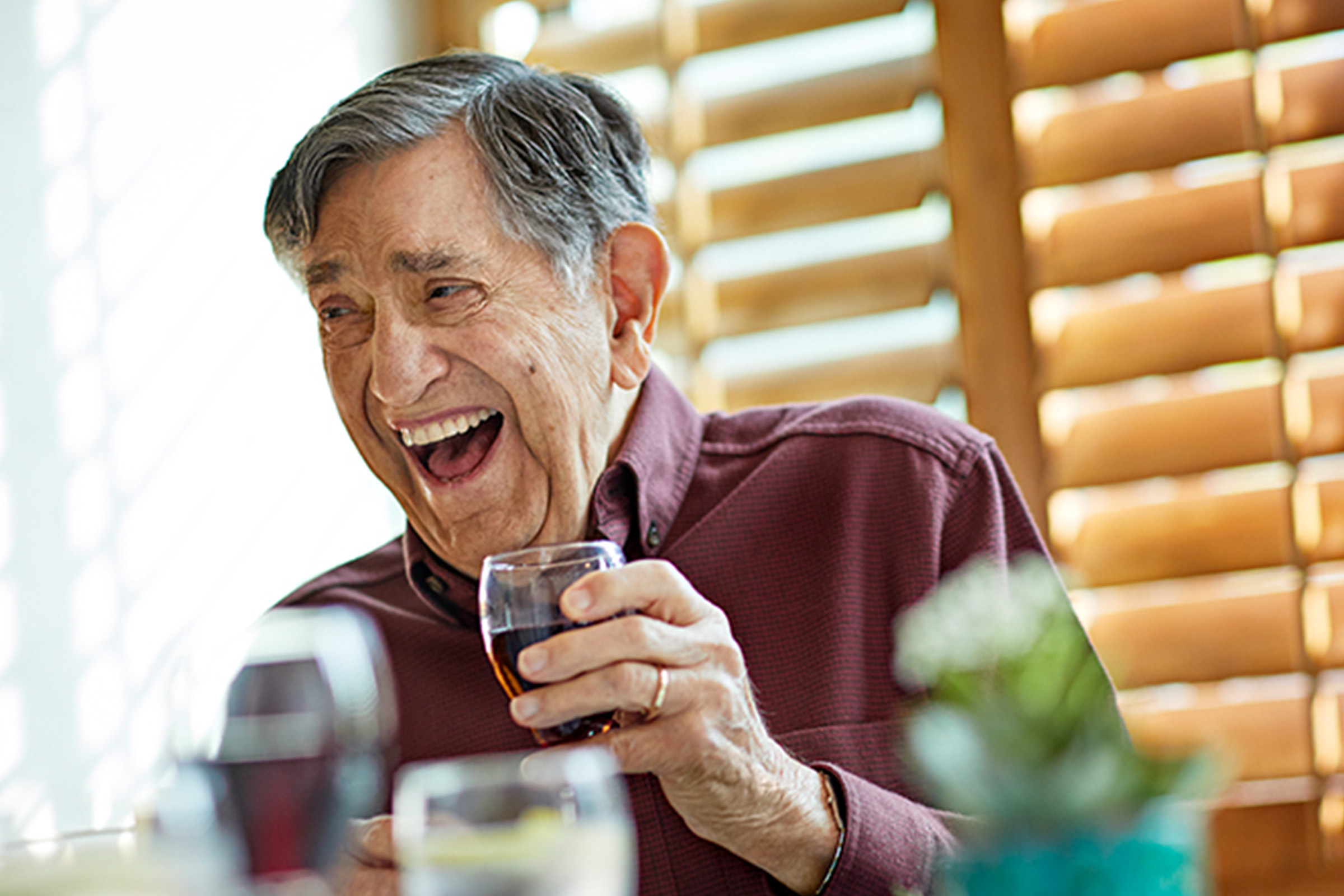 An elderly man in a maroon shirt sits by a window with wooden blinds, smiling and laughing while holding a glass of red wine. The foreground shows blurred drinks and a small plant.