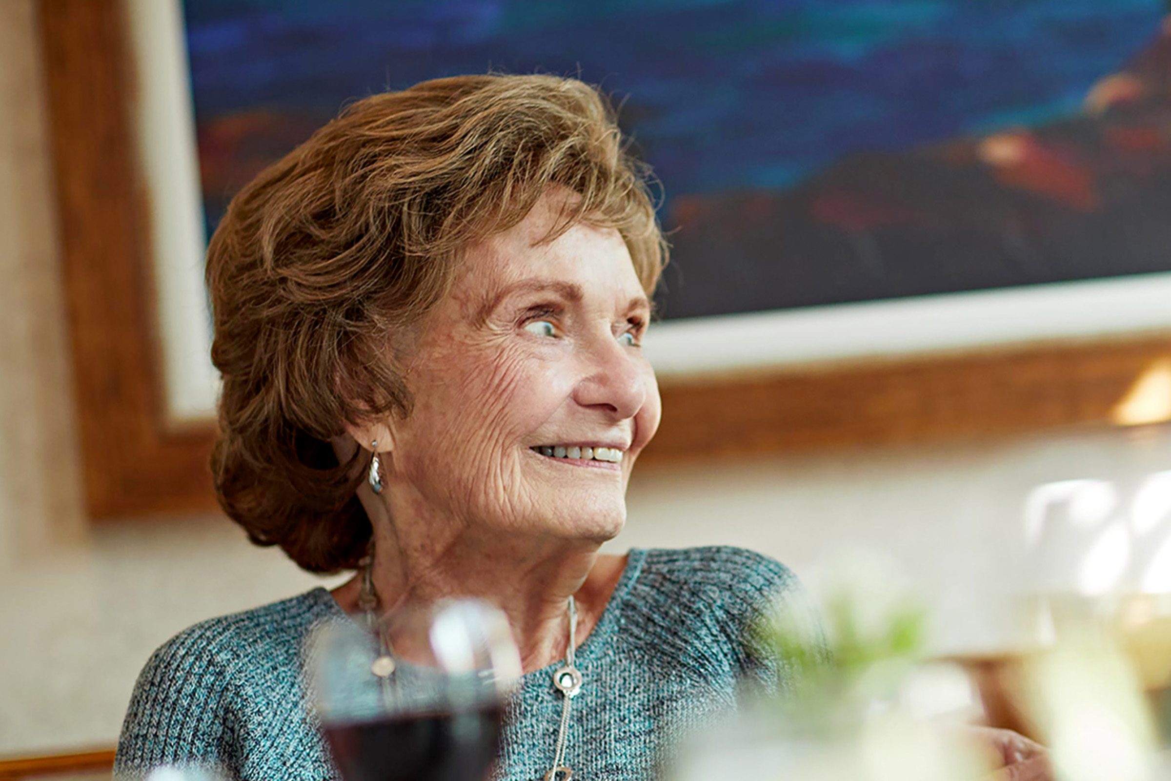 An elderly woman with short, wavy hair smiles while looking to the side. She is wearing a blue sweater and earrings, sitting indoors with blurred glasses and a plant in the foreground. A colorful painting is on the wall behind her.