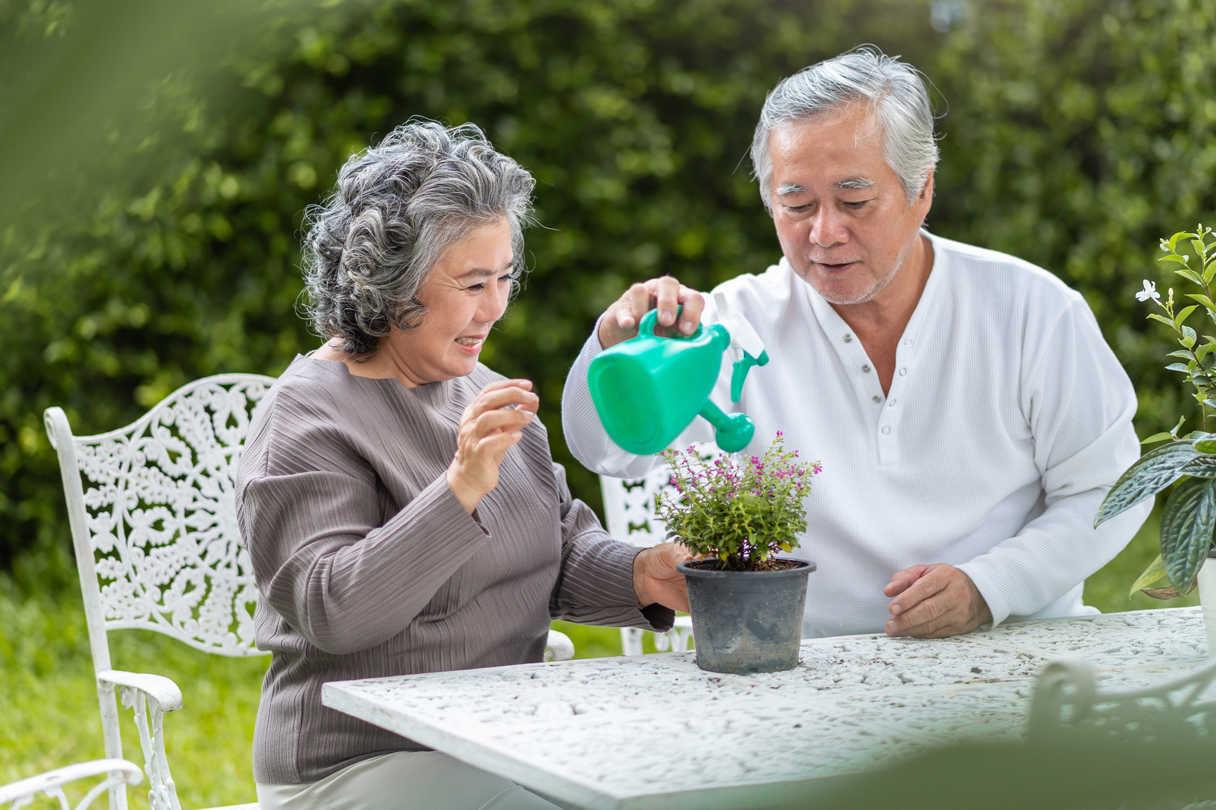 An elderly couple sits at a white outdoor table in a garden, smiling as the man waters a potted plant with a green watering can while the woman looks on and touches the plant gently.