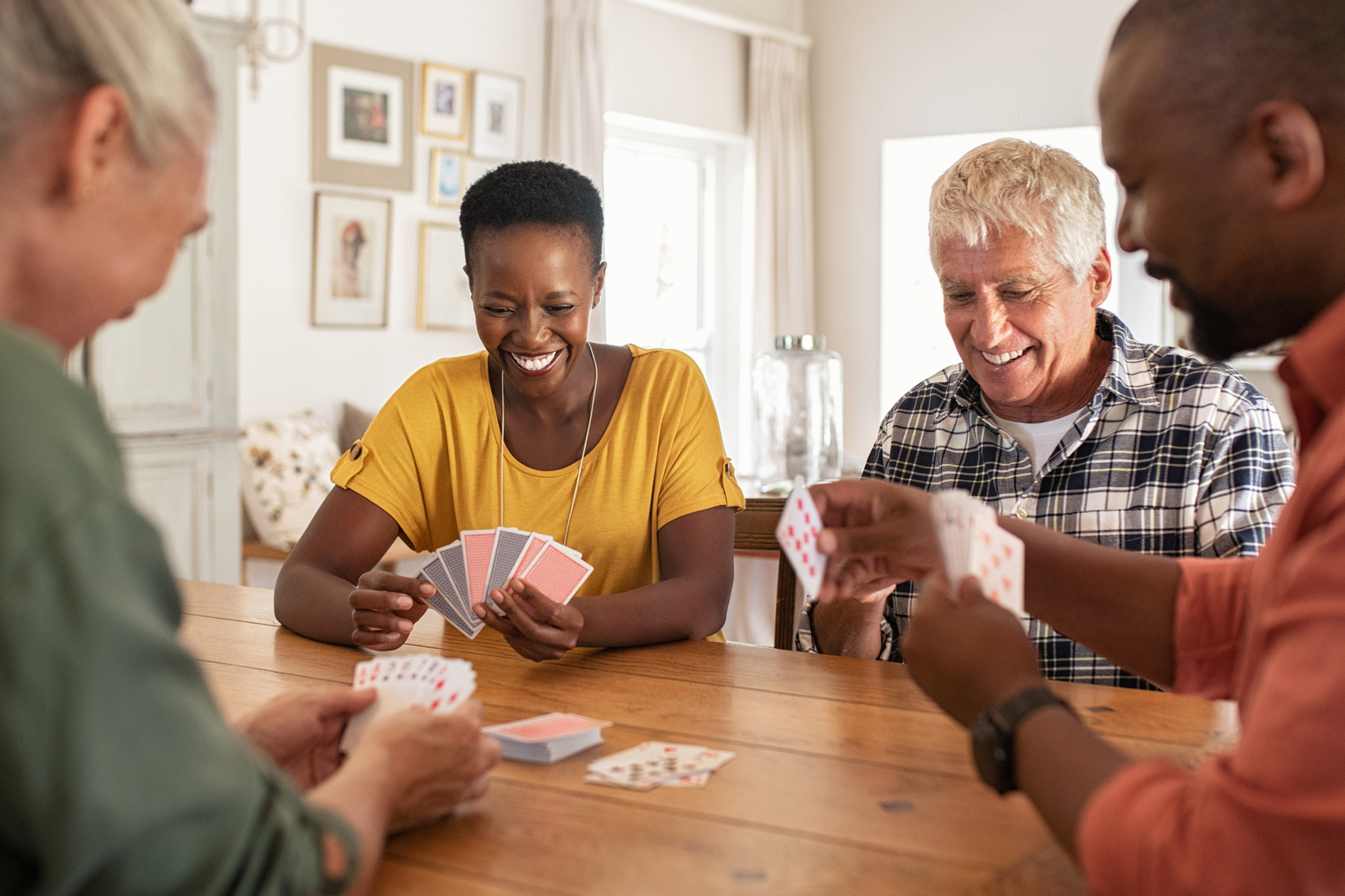 Four adults sit around a wooden table, smiling and laughing while playing cards together in a bright, cozy room. The mood is joyful and relaxed, and everyone is engaged in the game.