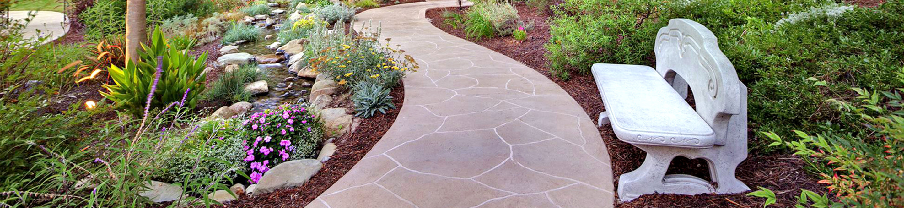 Curved stone pathway bordered by lush green plants, colorful flowers, and rocks leads through a garden with a decorative white bench on the right side.