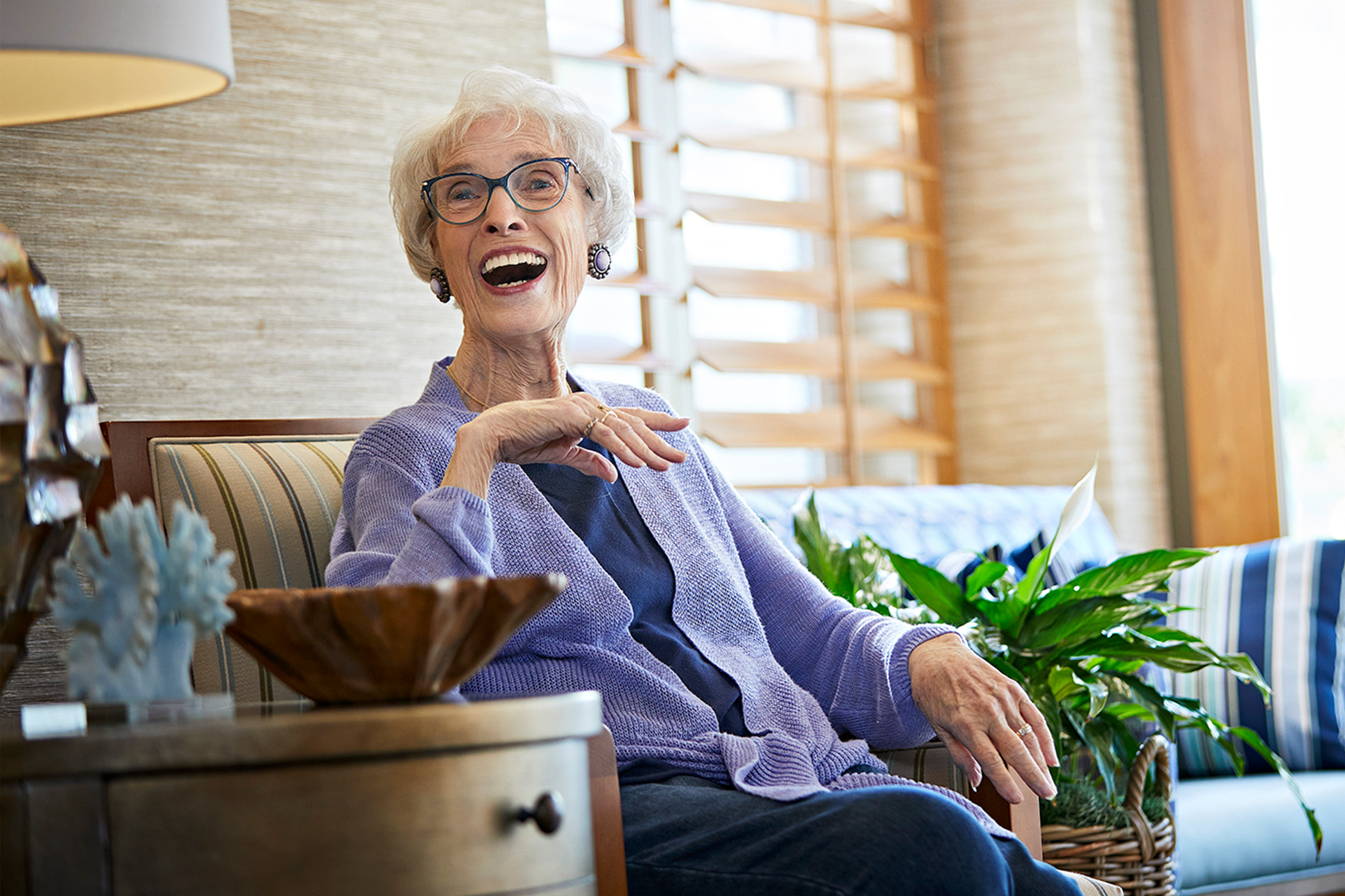 An elderly woman with short white hair and glasses sits smiling and laughing on a striped armchair in a bright, cozy living room, with a wooden bowl and a green plant nearby.
