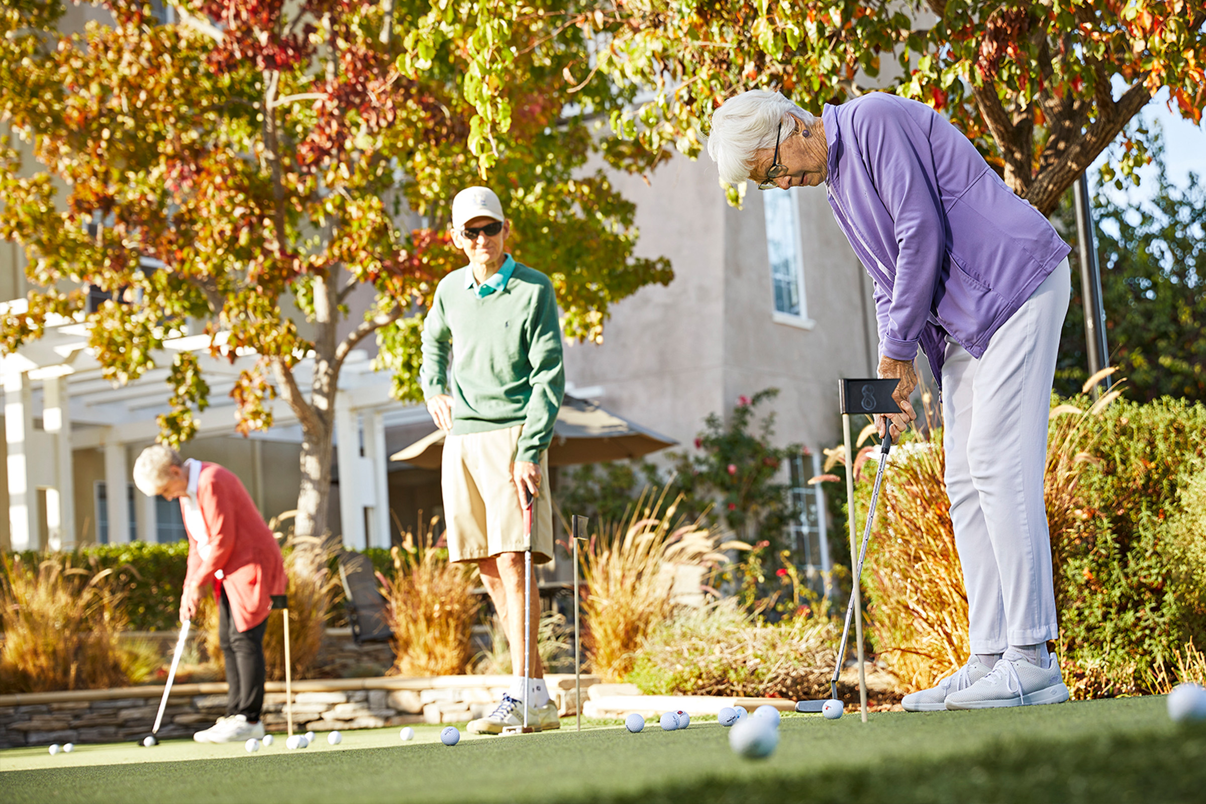 Three seniors play mini-golf outside on a sunny day; one woman is putting while another woman and a man watch in the background, surrounded by trees and a building.