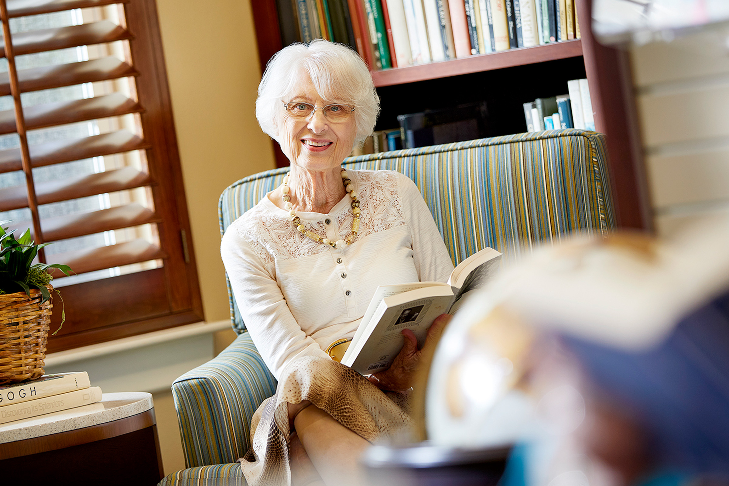 An elderly woman with short white hair and glasses sits in a striped armchair, smiling while holding an open book. Behind her are bookshelves filled with books, and a window with wooden blinds lets in natural light.